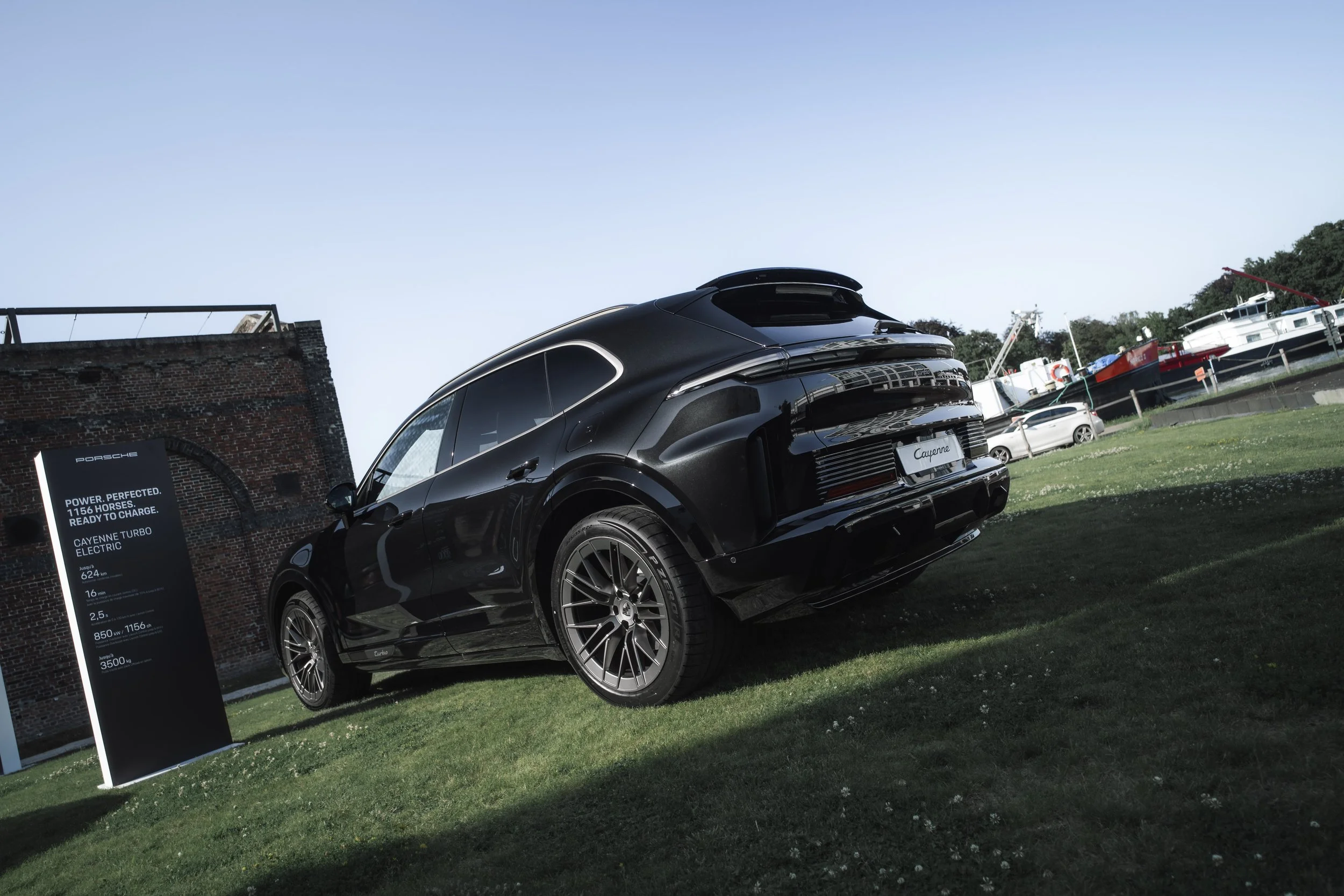 A black Porsche Cayenne Turbo electric SUV parked on grass near boats docked at a marina, with a sign displaying vehicle specifications nearby.