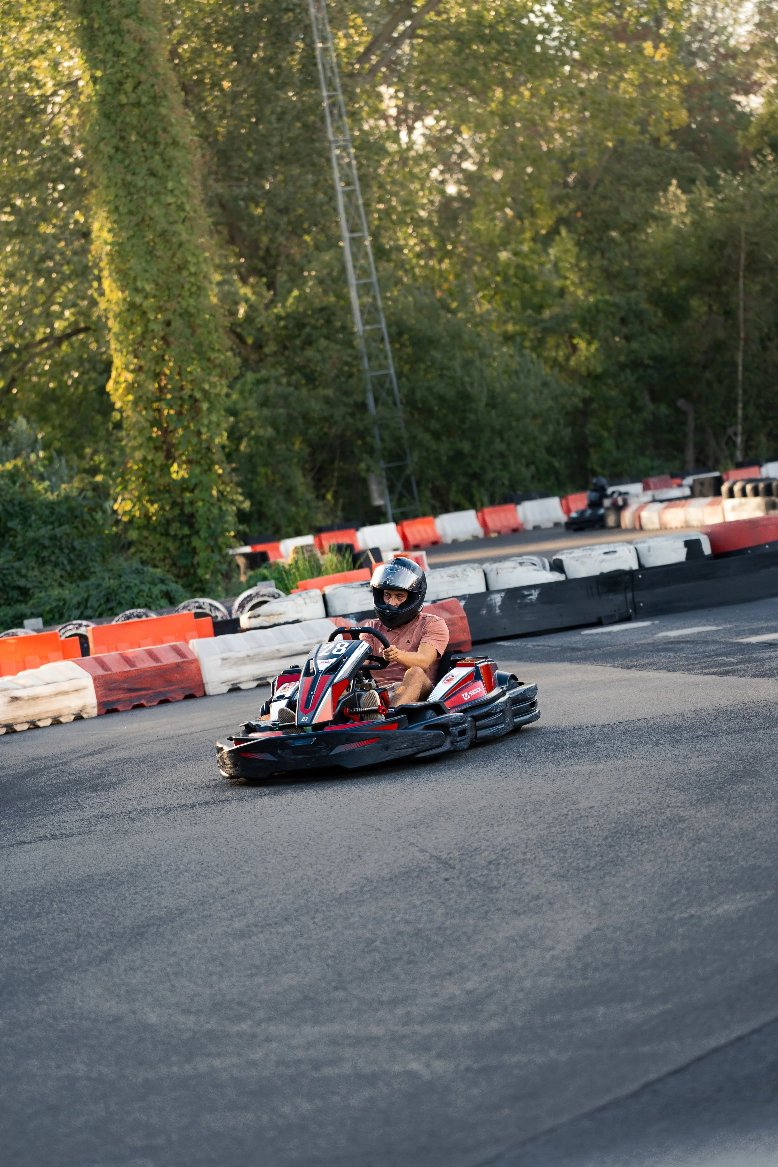 Person in a black helmet driving a red and black go-kart on a race track, surrounded by red and white barriers, with green trees in the background.