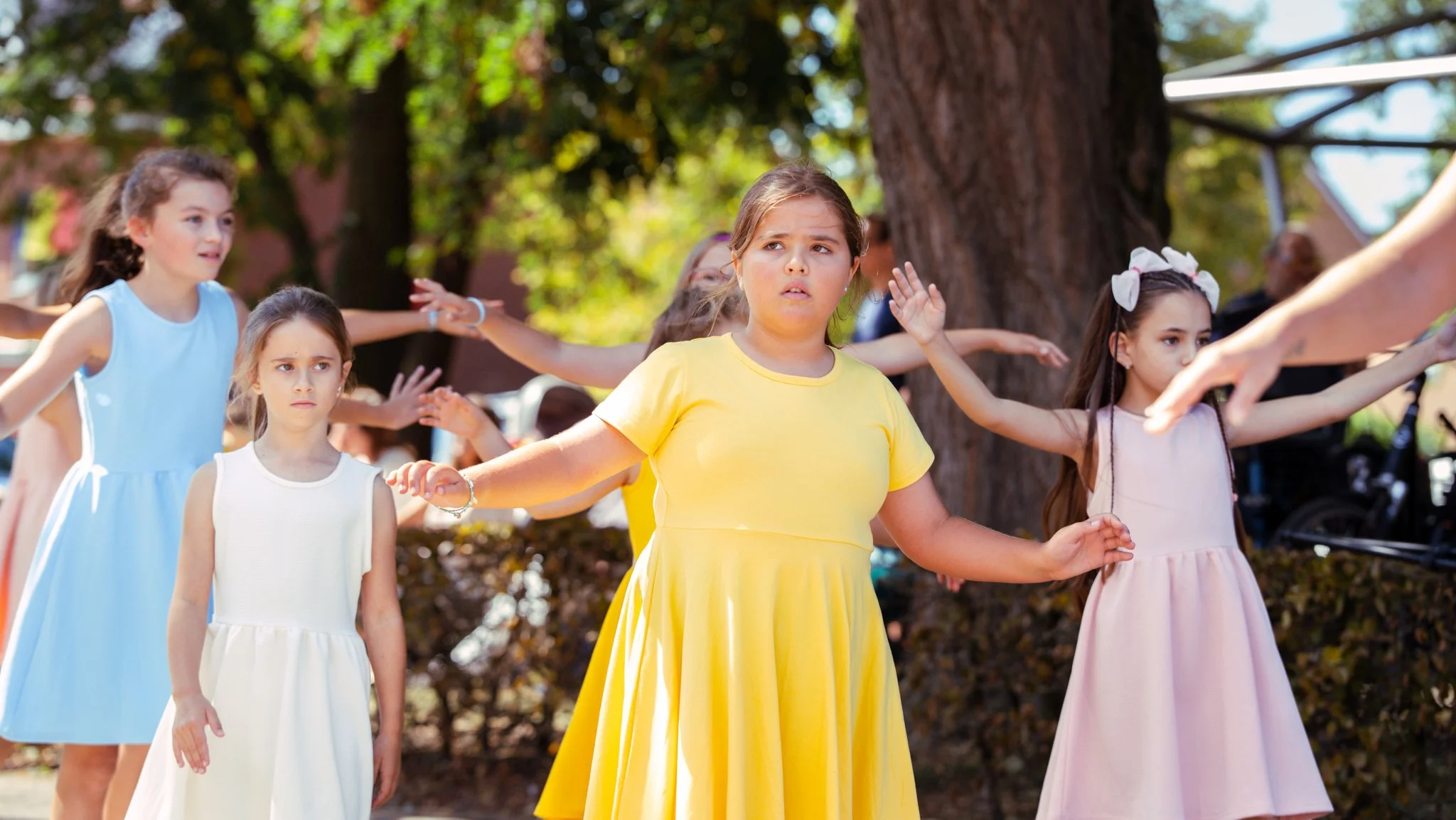 A group of young girls participating in an outdoor activity, standing with arms extended, with trees and sunlight in the background.