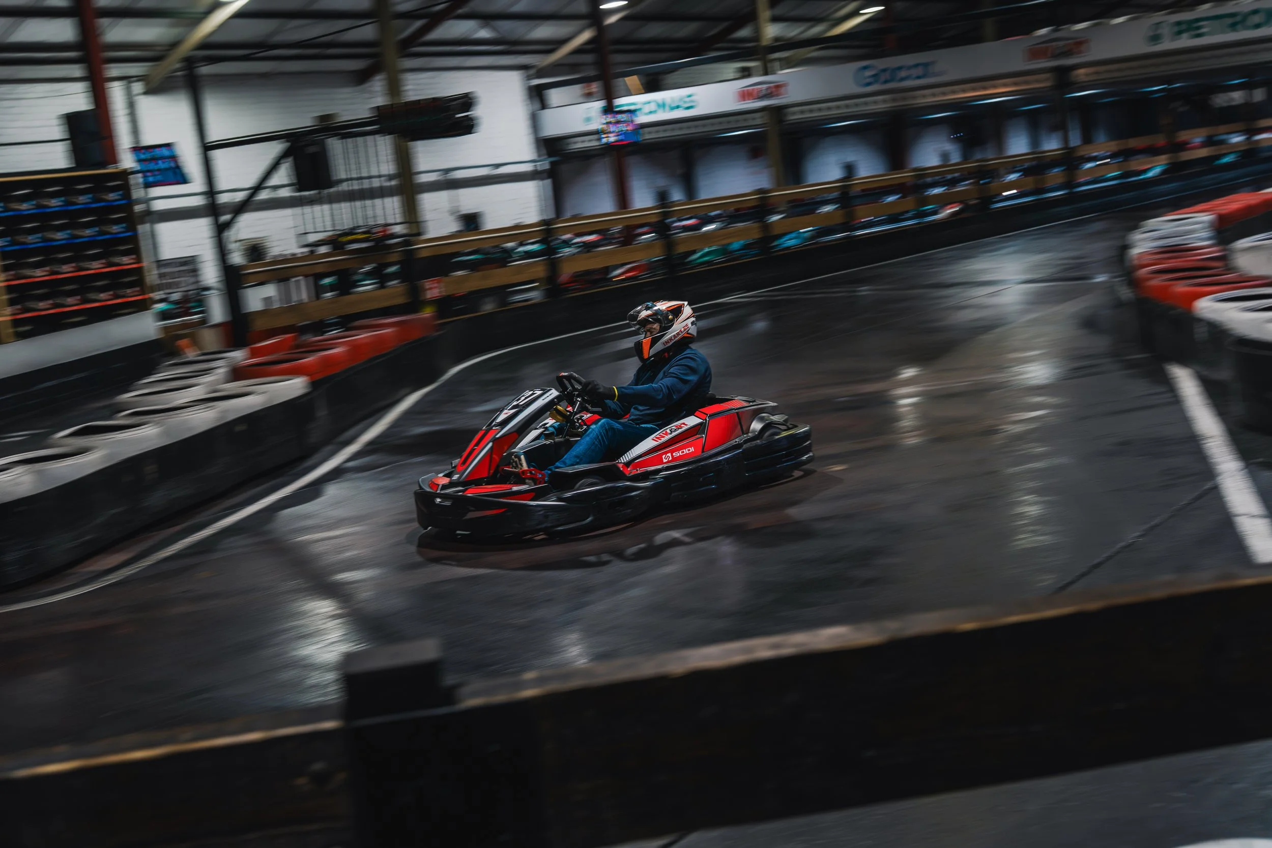 A person in a racing helmet and blue jacket driving a go-kart on an indoor race track with black and white barriers and orange and white tires.