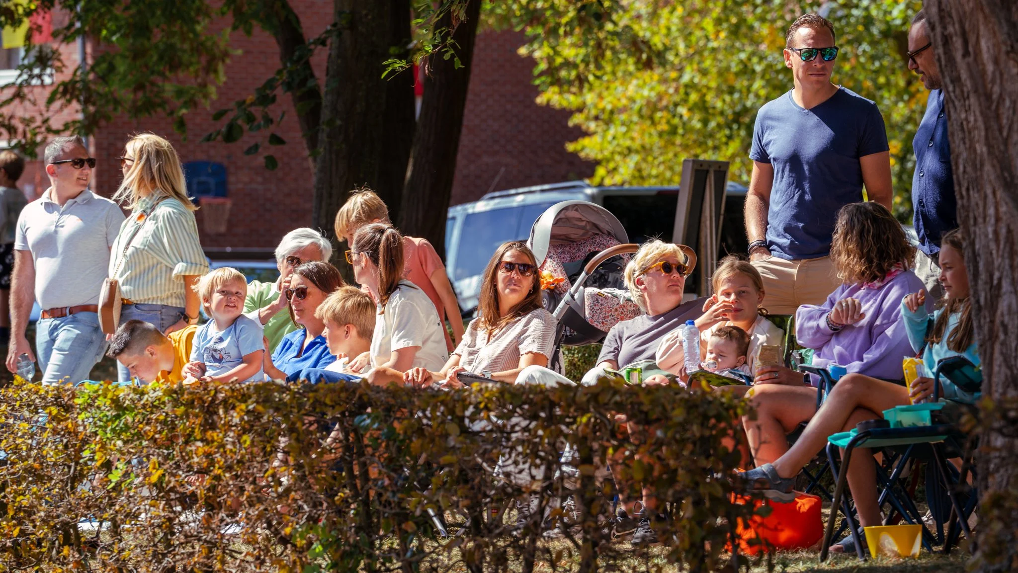 Group of people, including children and adults, sitting and standing outdoors on a sunny day in a park, with trees and a brick building in the background.