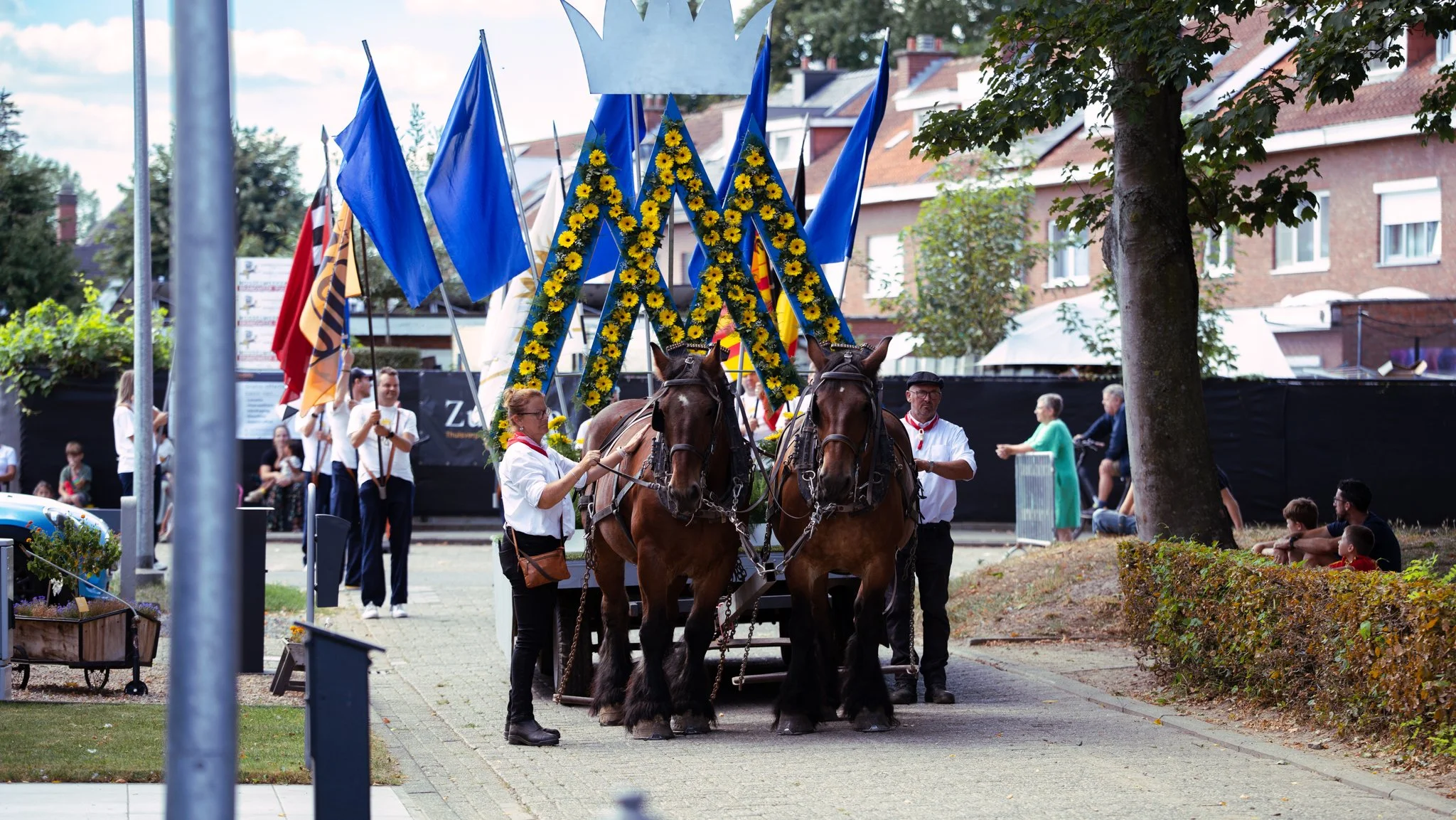 A parade float decorated with large, yellow flowers and blue flags being pulled by two horses. People are walking alongside and in the background are spectators and residential buildings.