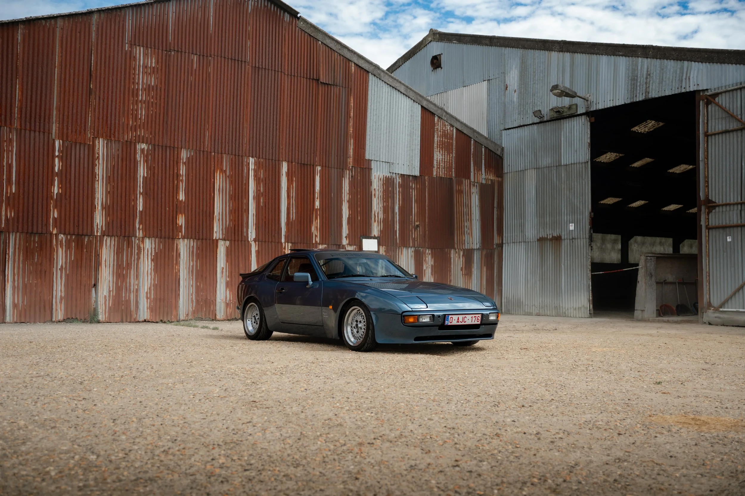 A dark gray vintage sports car with a European license plate parked in front of large rusted metal warehouse buildings under a partly cloudy sky.