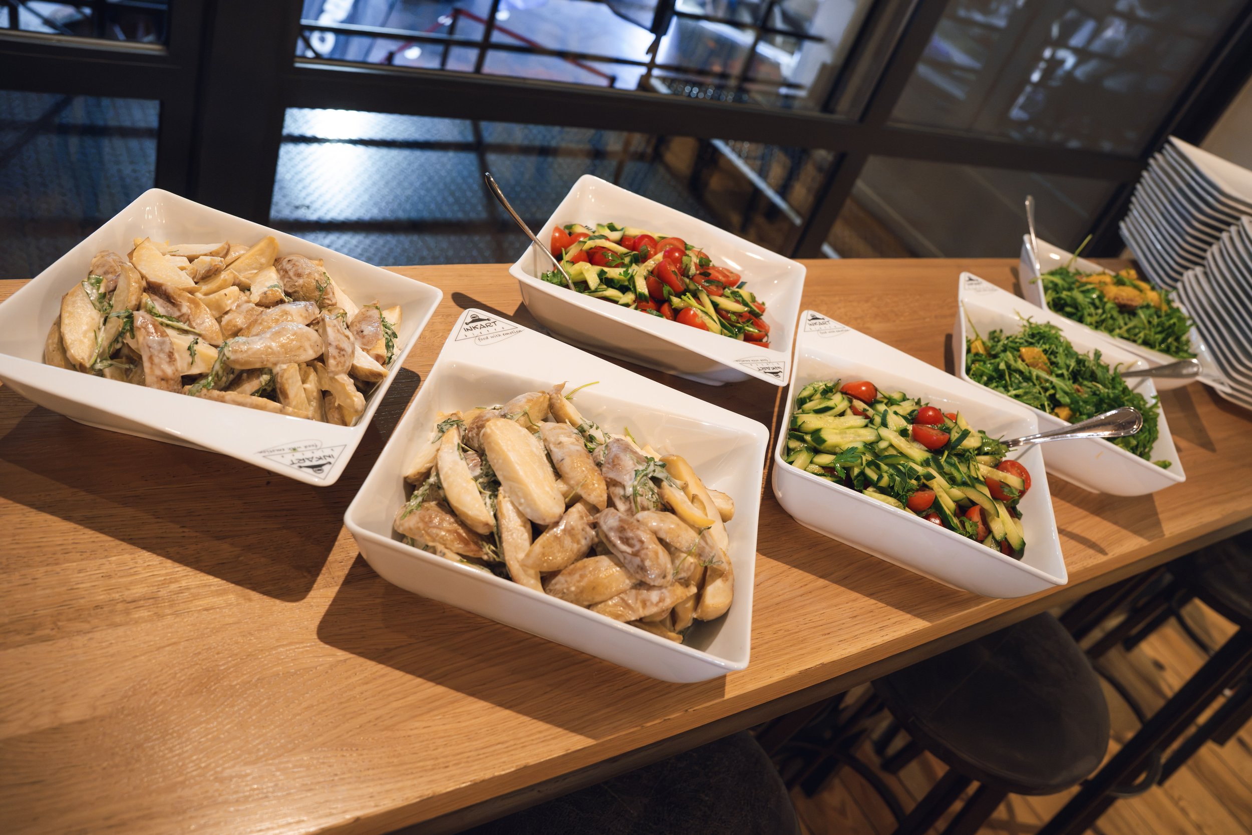 Assorted salads in white bowls on a wooden table at a restaurant, with a window with black frames in the background.