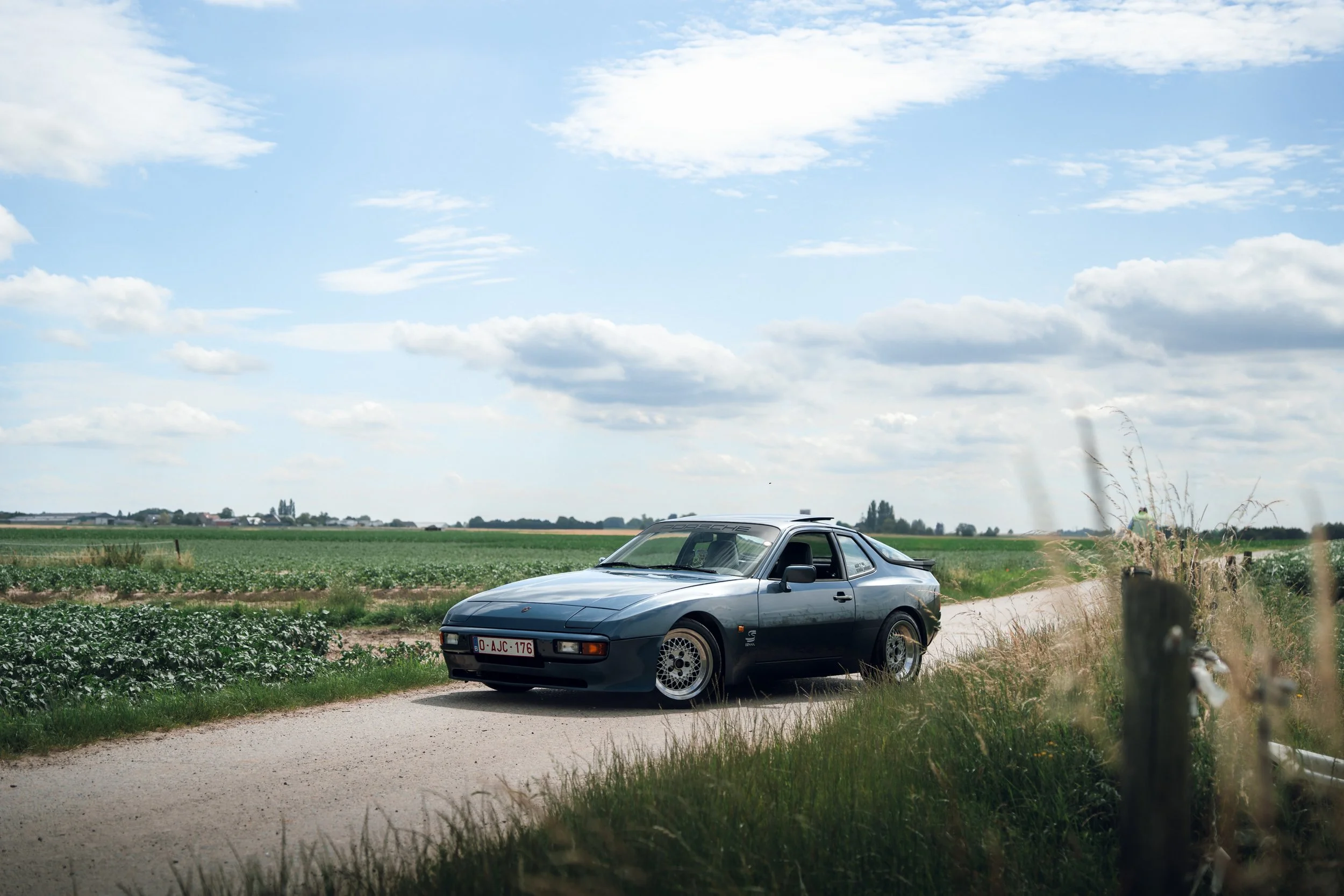 A classic gray sports car parked on a rural dirt road near green fields and overcast sky.