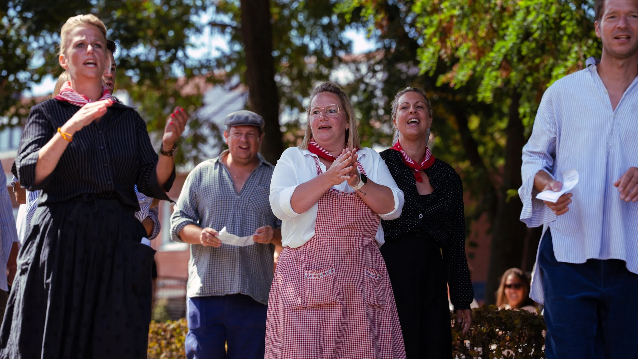 Group of people outdoors, some wearing checkered shirts and aprons, possibly involved in a event or celebration, with a background of trees and a building.