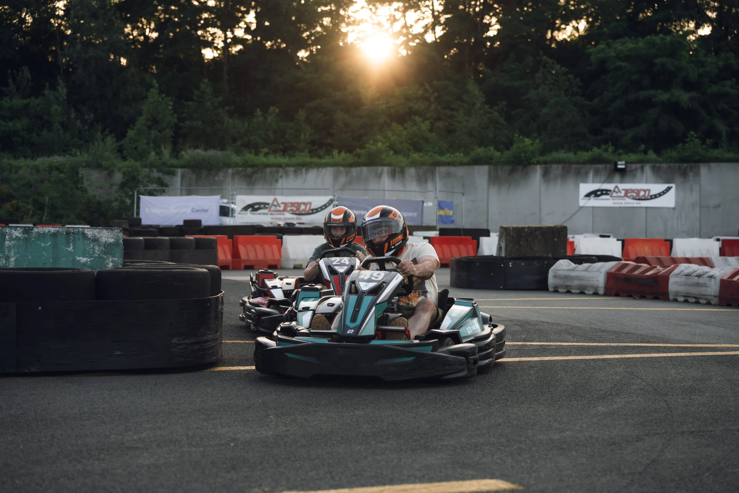 Two individuals wearing helmets and sunglasses riding electric go-karts on an outdoor track during sunset, with tires and barriers around the course and trees in the background.