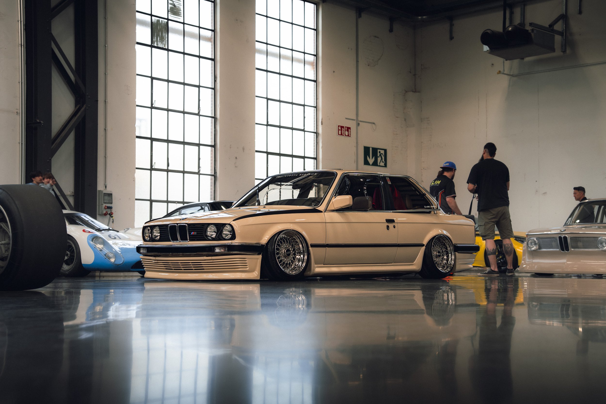 Classic beige BMW car with black wheels inside a spacious industrial-style building, with other vintage cars and several people in the background.