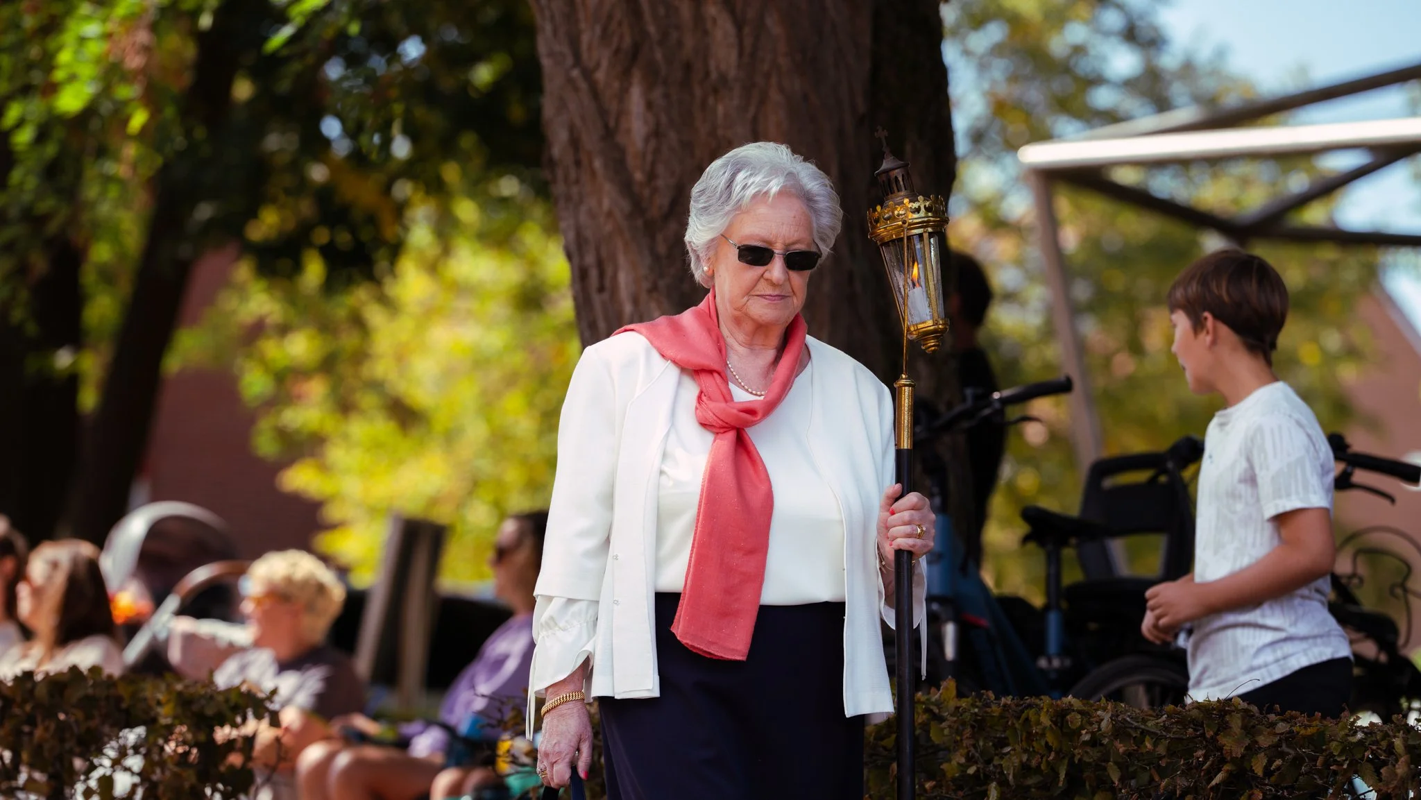 An elderly woman with white hair and glasses stands outdoors holding a lantern, wearing a white jacket with a pink scarf, with a young boy in a white shirt nearby, in a park-like setting with trees and blurred people in the background.