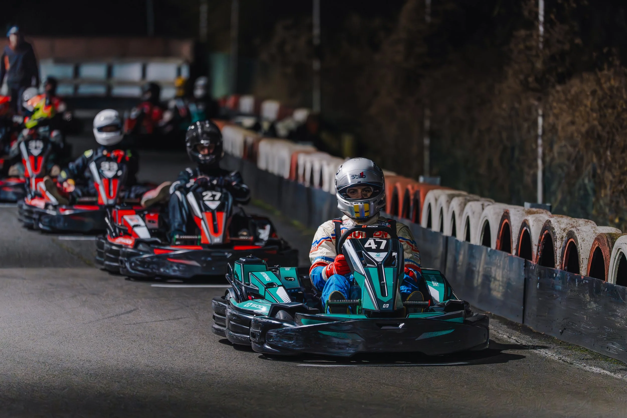 Go-kart racers in helmets and racing suits driving in line along a track with safety barriers and a forested background.
