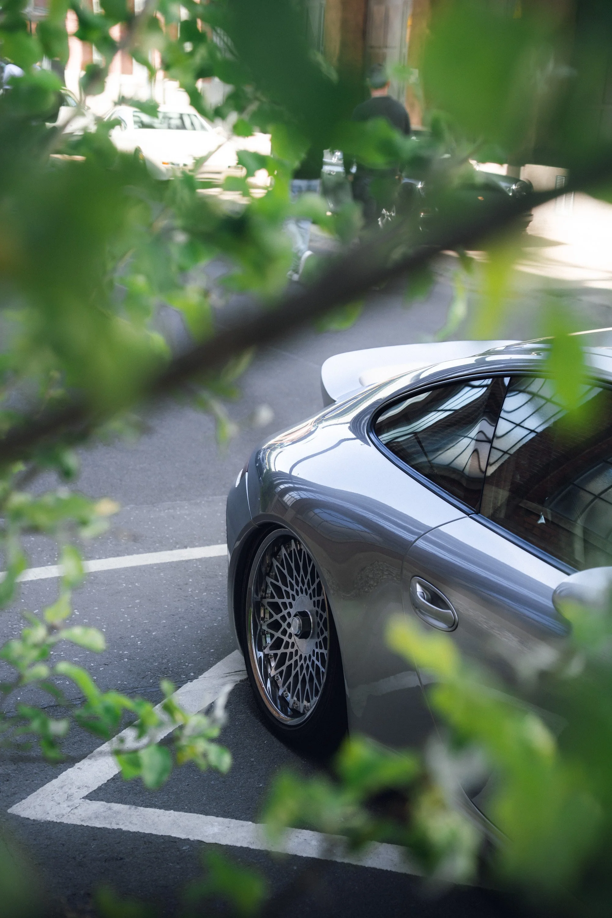 The image shows a silver car parked in a parking lot, partially obscured by green leaves and foliage in the foreground. Two people are walking along the sidewalk in the background, and several other parked cars are visible along the street.