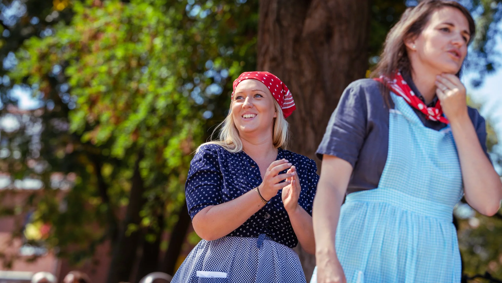 Two women in vintage-style clothing and headscarves standing outdoors near a tree, smiling and interacting.