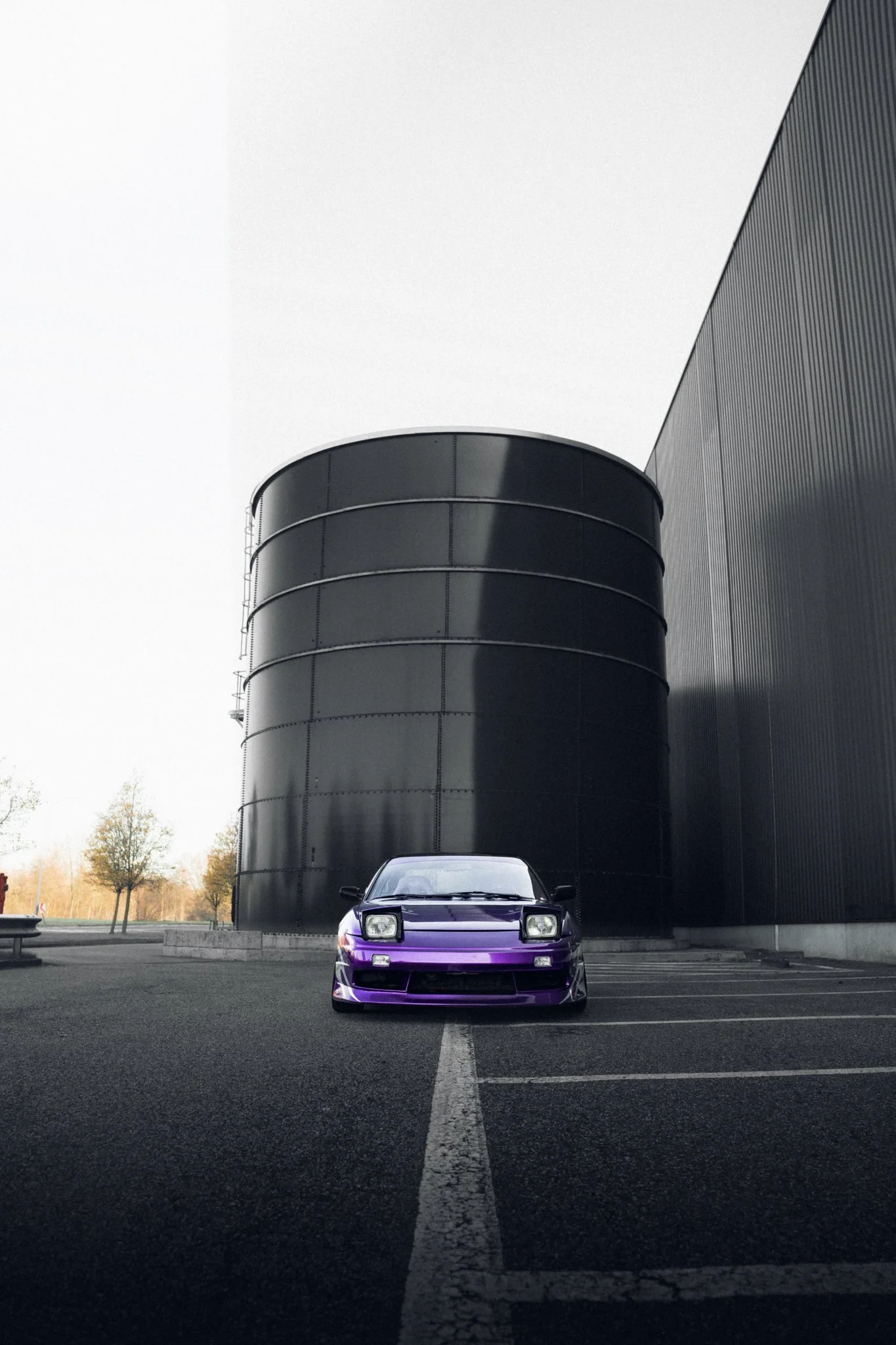 A purple sports car parked in an empty parking lot in front of a large black industrial building with a curved wall.
