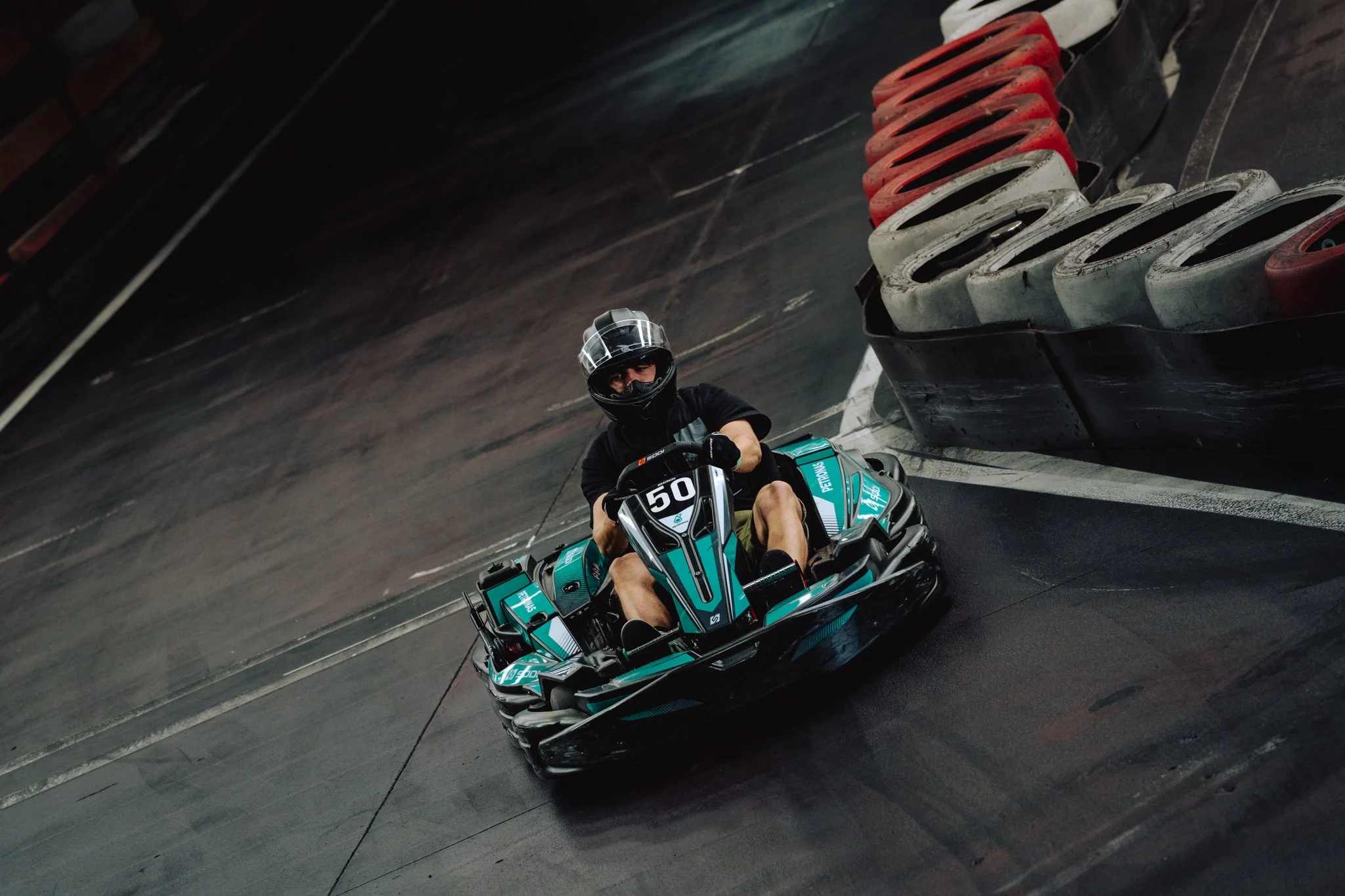 A person wearing a black helmet and black clothing driving a blue and black go-kart on an indoor race track.