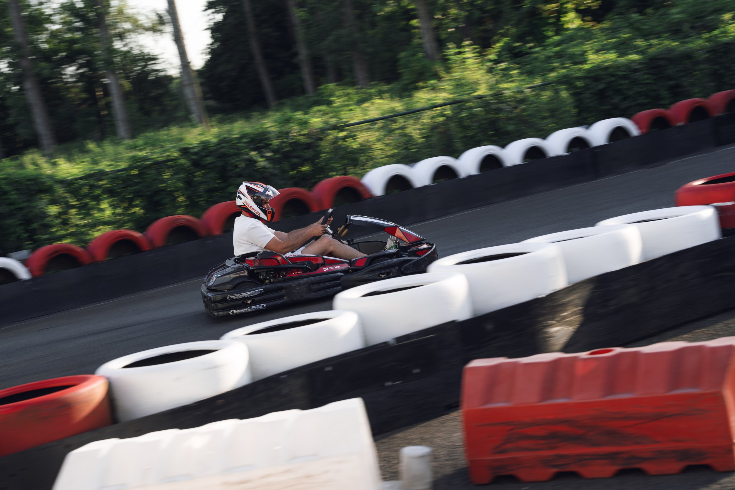 A person driving a go-kart on an outdoor race track with red and white barriers, surrounded by green trees.