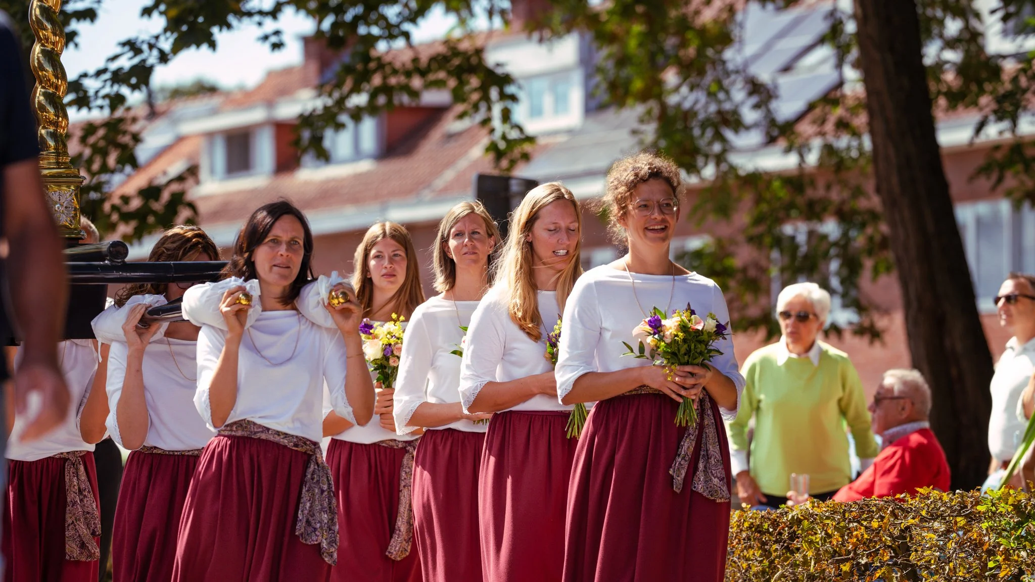 Group of women in traditional attire participating in a ceremonial event outdoors, some holding flowers, with onlookers and trees in the background.