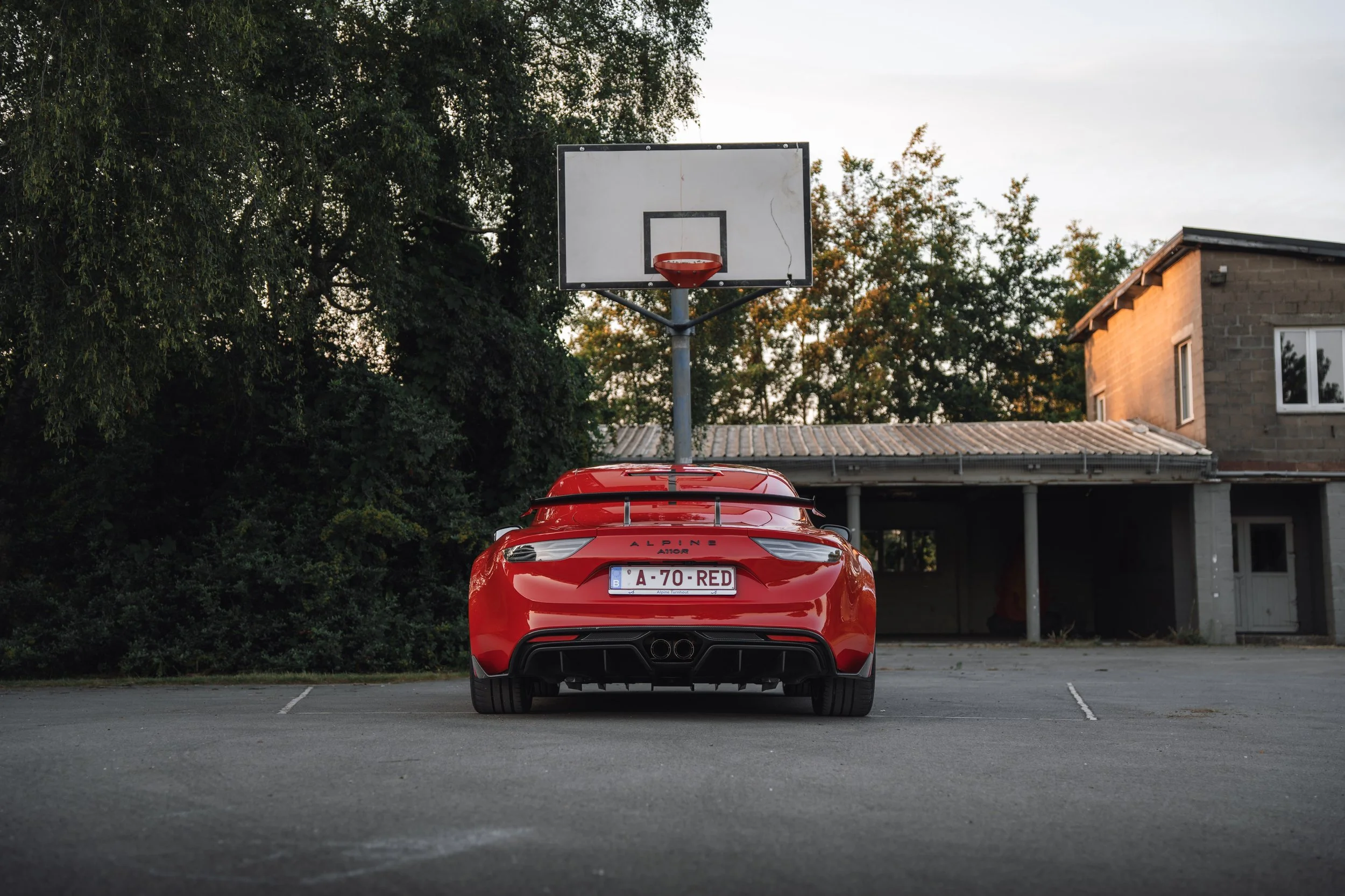 Red sports car parked in an empty parking lot with a basketball hoop and residential building in the background during daylight.