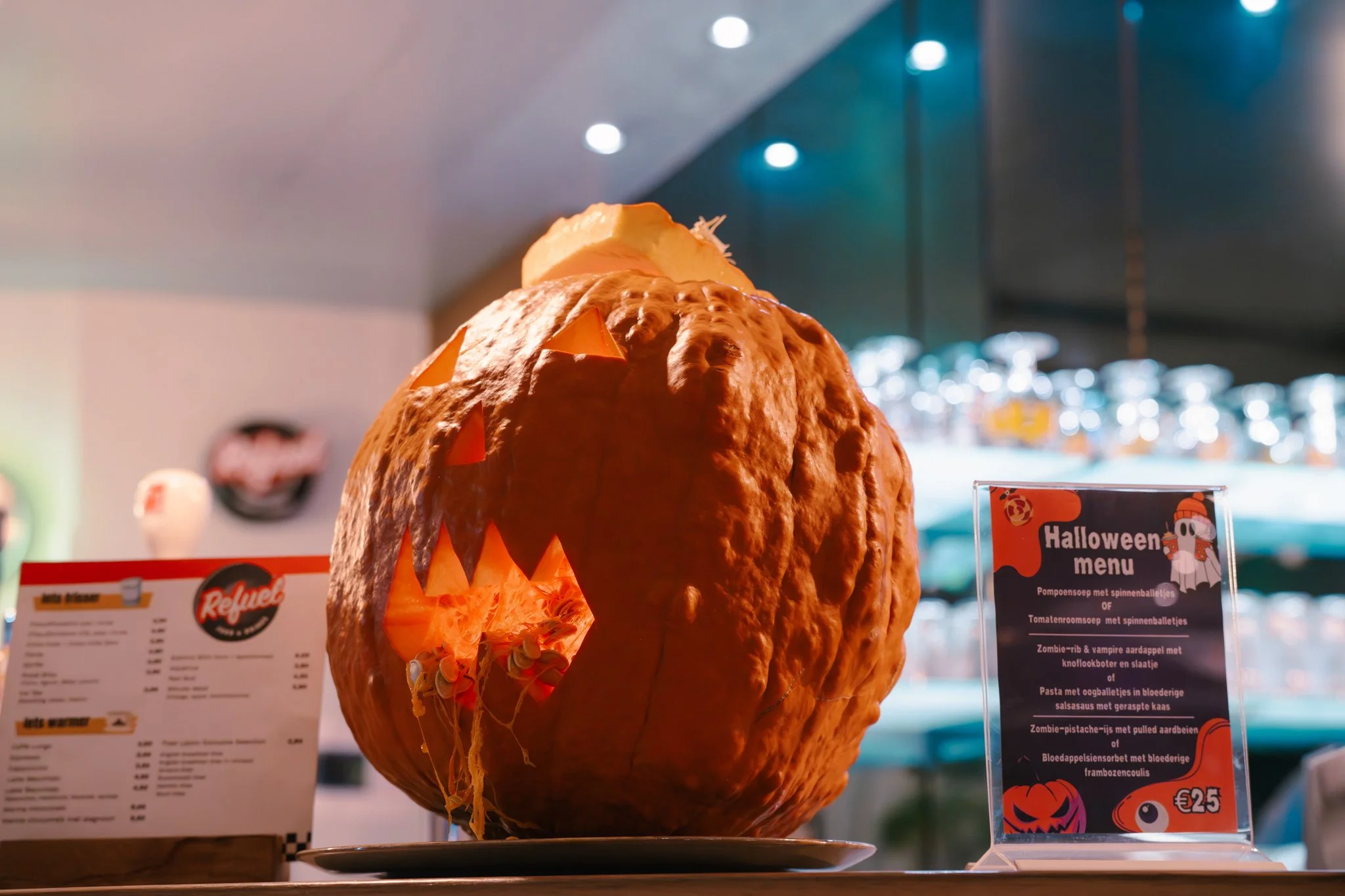 A carved pumpkin with a spooky face, illuminated from inside, displayed on a table at a Halloween event with Halloween-themed menus nearby.