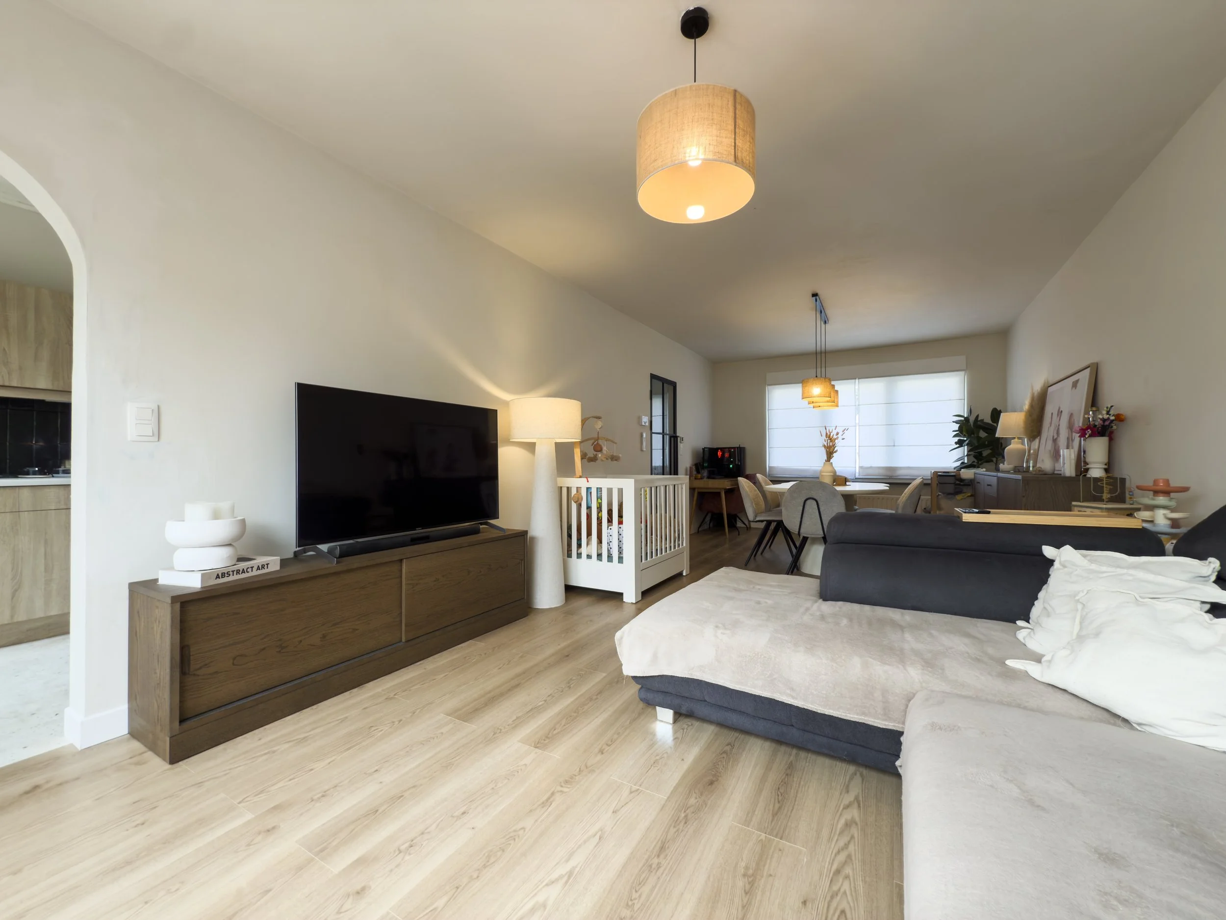 Living room with a beige sectional sofa, a TV on a wooden stand, a white floor lamp, and a crib, with a dining area in the background featuring a round table, chairs, and pendant lights.