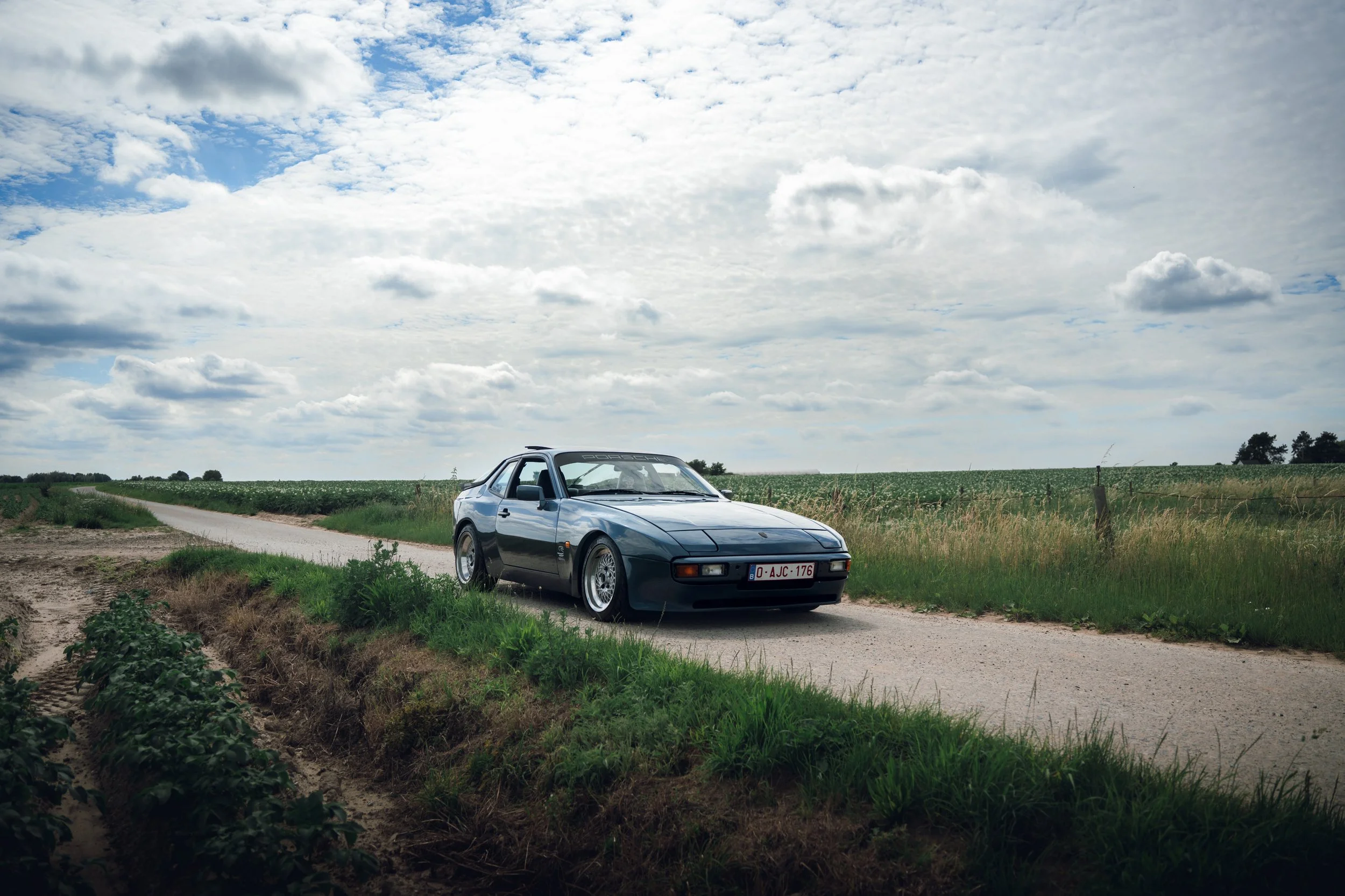 A dark gray vintage sports car parked on a rural dirt road with green fields and cloudy sky in the background.
