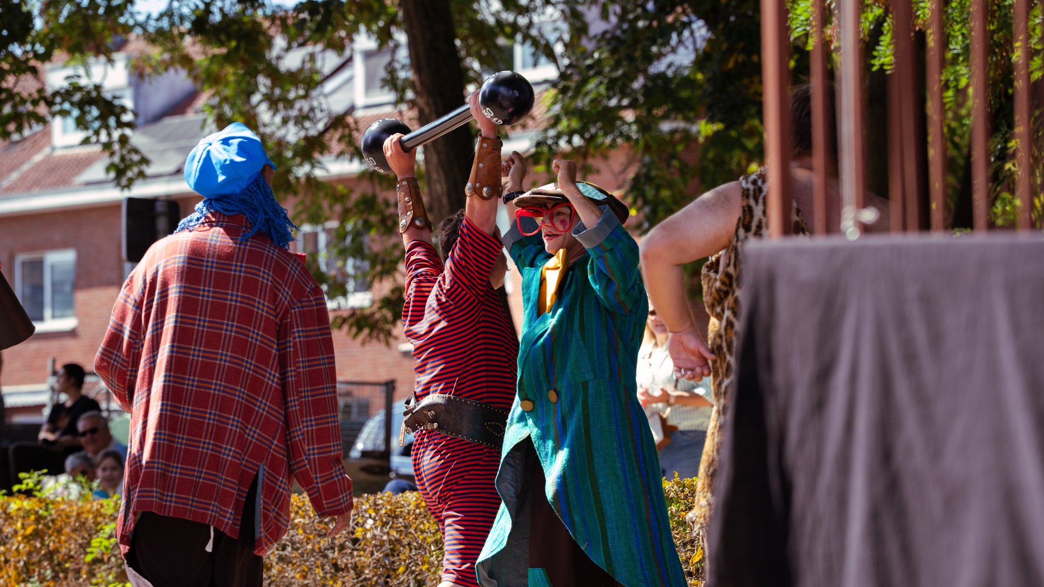 Group of people dressed as clowns and performers having fun outdoors, with one in a colorful striped coat and large glasses, holding a dumbbell-shaped object, at a festival or parade.