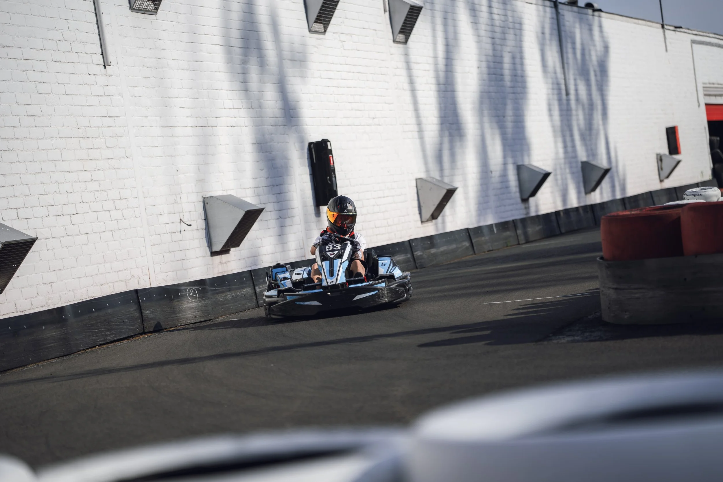 A person wearing a helmet riding a go-kart on an outdoor racing track, with a white brick wall in the background and red barriers on the sides of the track.