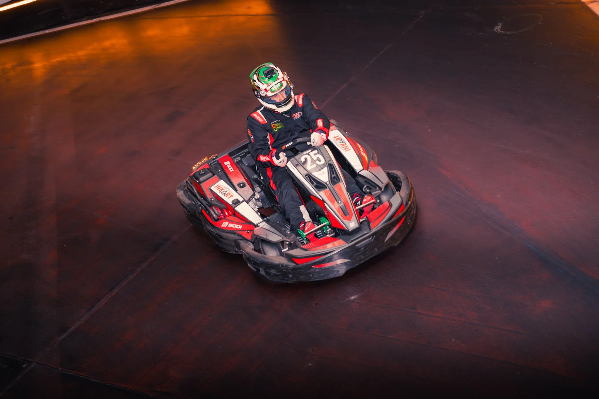 A person wearing a racing suit and helmet driving a red and black bumper car on an indoor ice rink or skating rink.