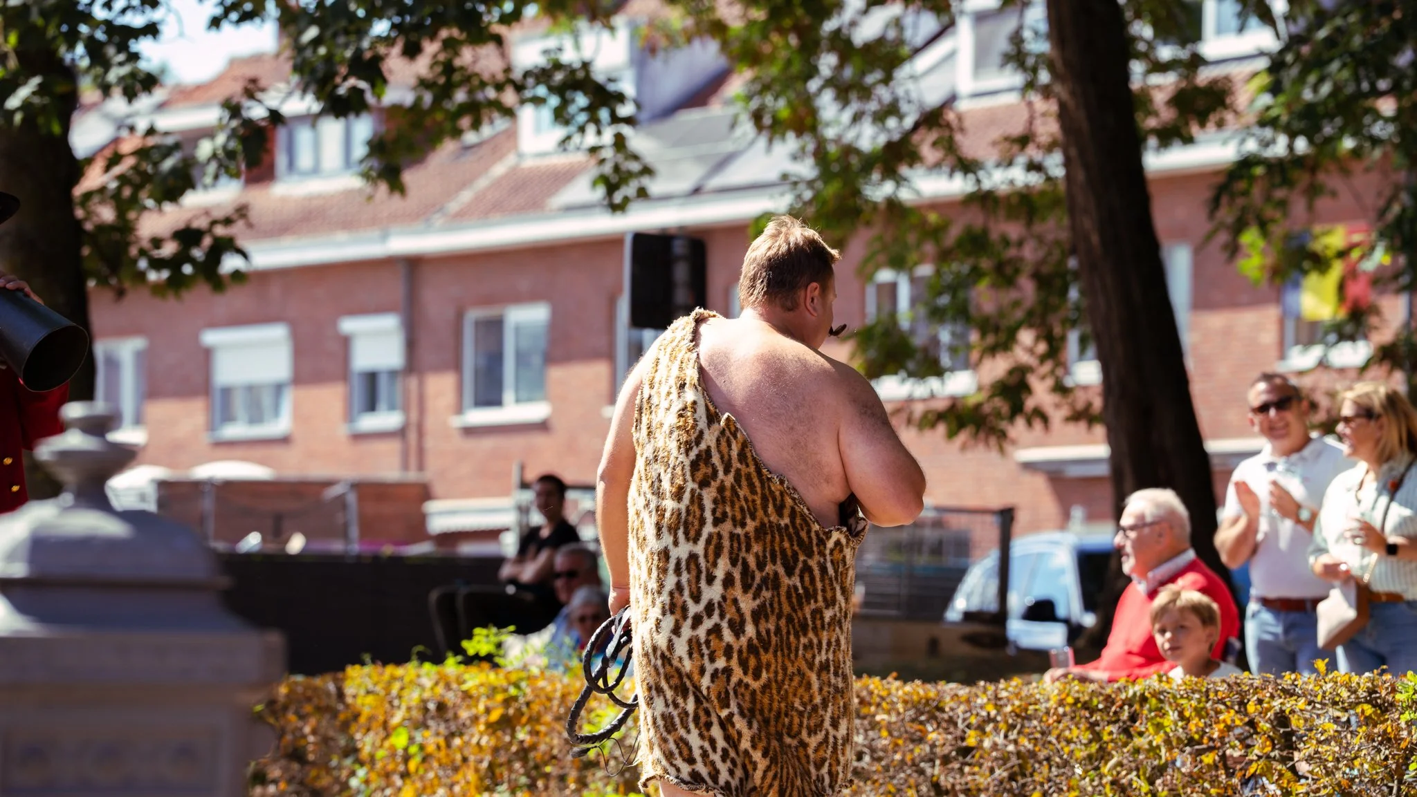 Man dressed in a leopard print costume performing as a street entertainer during a sunny outdoor event, with people watching and smiling in the background.
