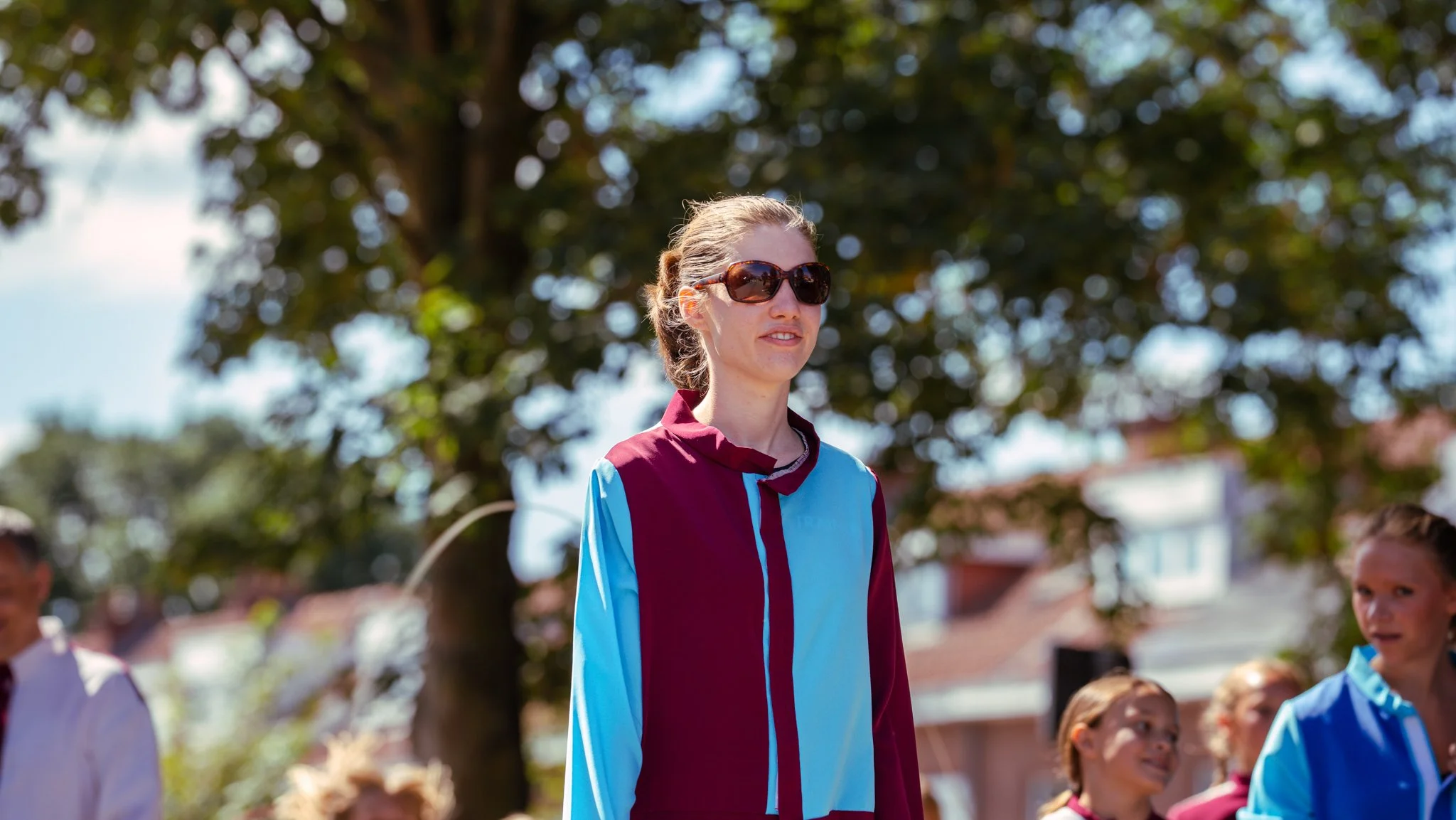A woman with sunglasses standing outdoors among children, with trees and buildings in the background.