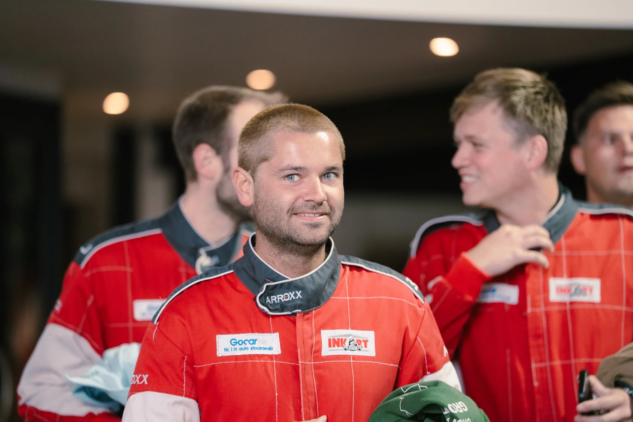 Group of male race car drivers wearing red racing suits, standing indoors, with some smiling and talking.