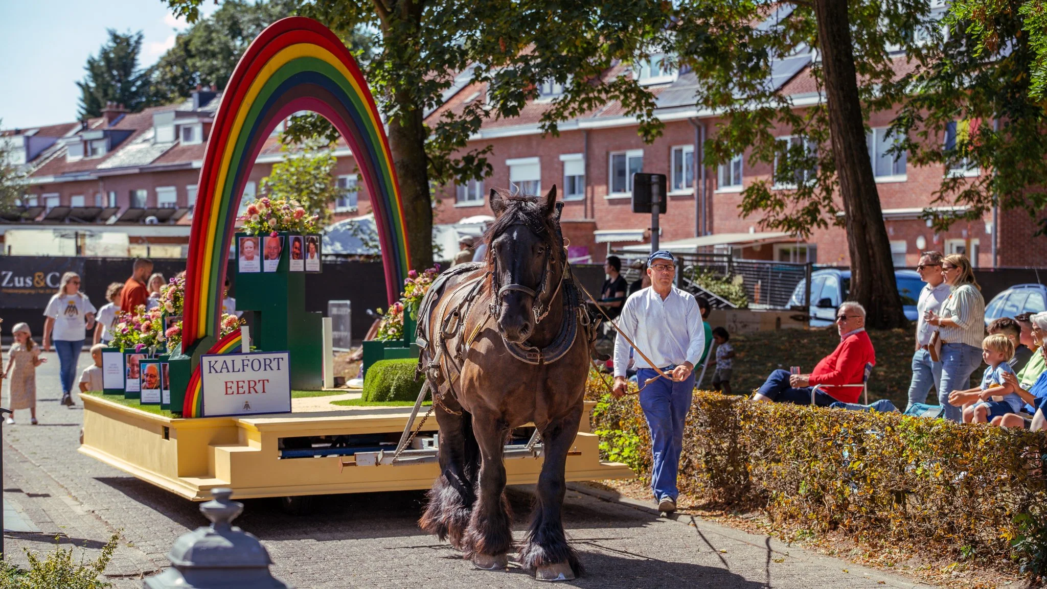 A decorated parade float featuring a rainbow sculpture, with a horse pulling it, and spectators watching along the street.
