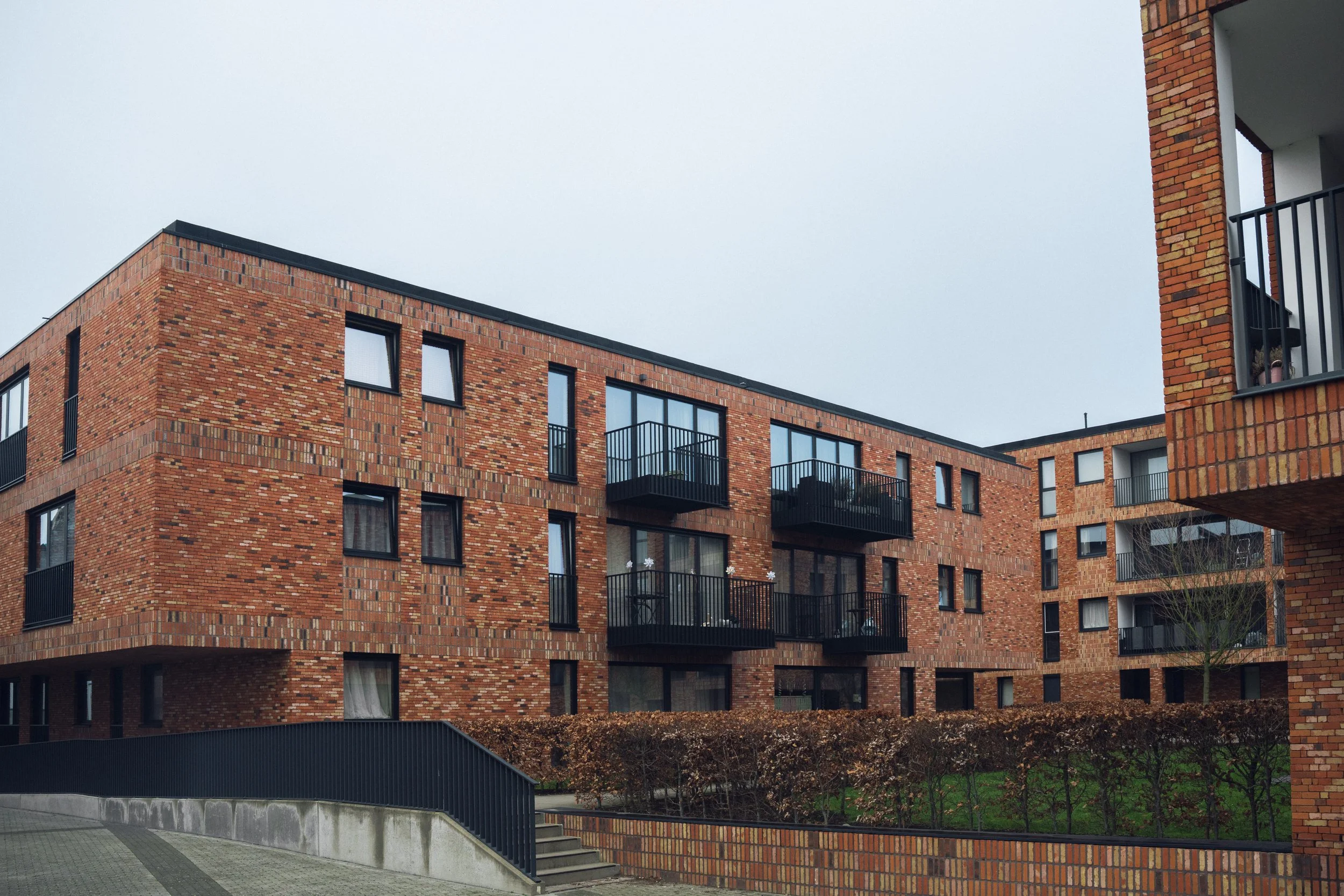 Brick apartment building with black balconies and several windows, some with curtains, on a cloudy day.
