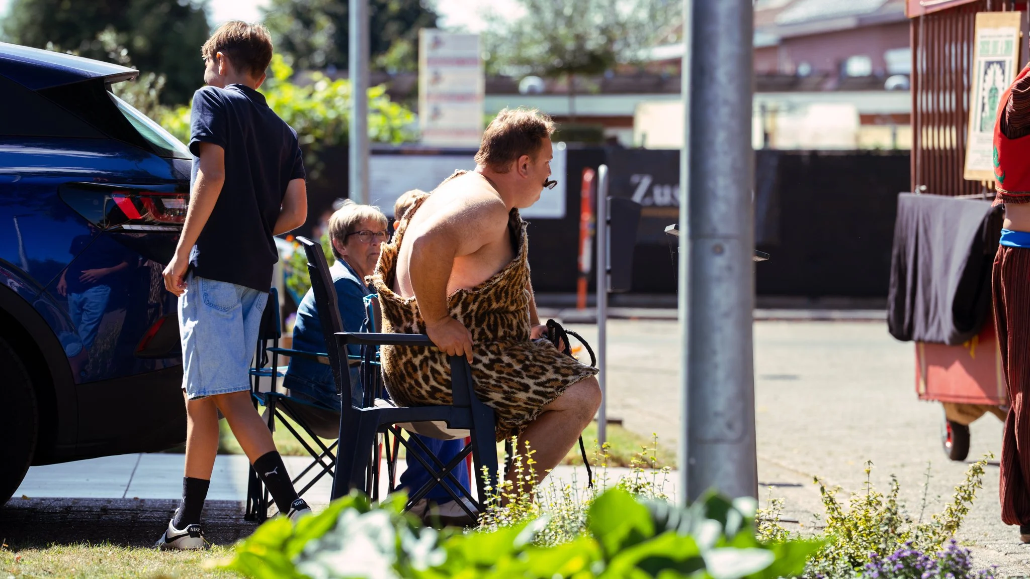 Man dressed in a leopard print costume sitting on a folding chair at a sidewalk event, with a woman wearing glasses and a backpack sitting behind him, and children walking by.
