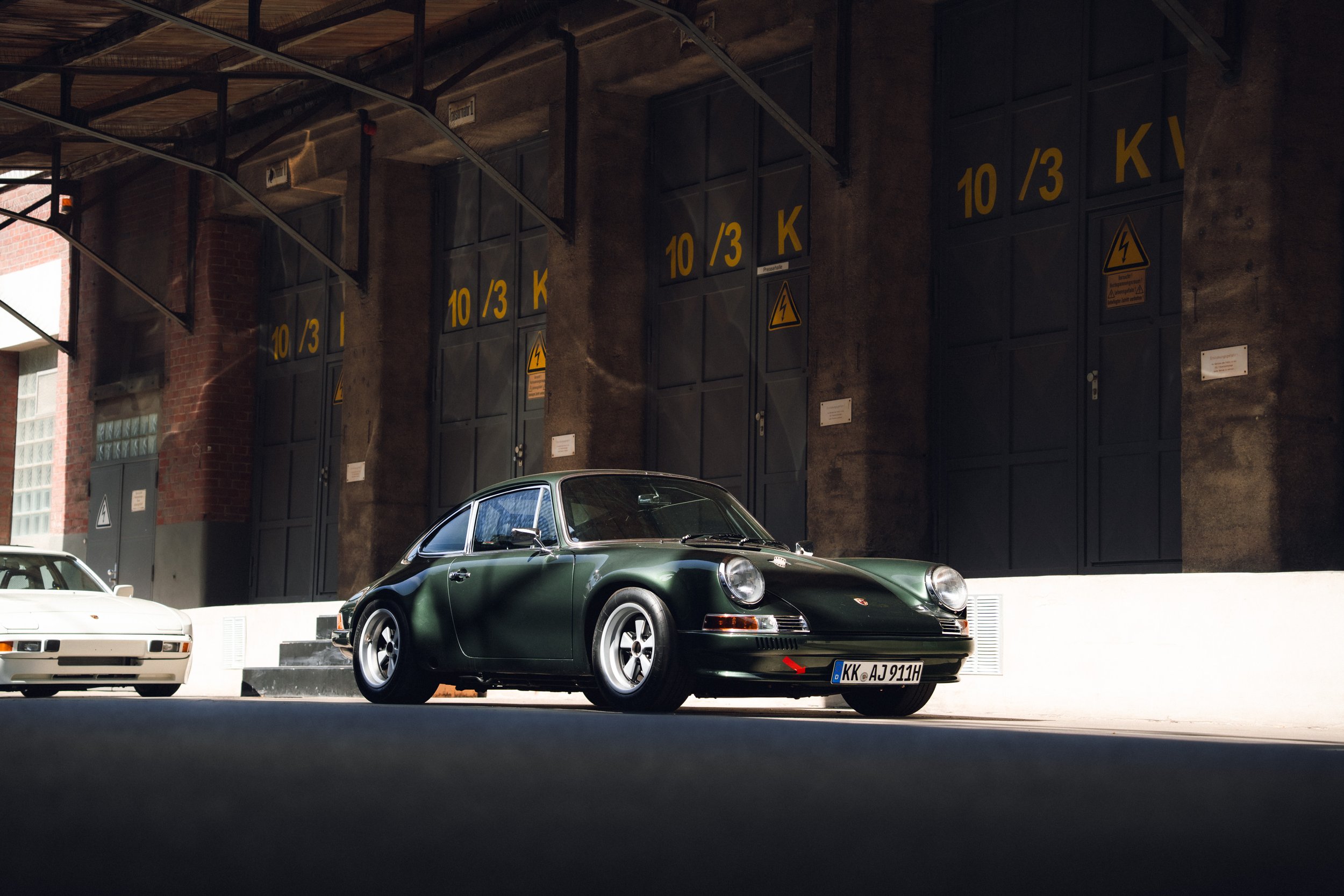 A dark green vintage Porsche sports car parked on a street in front of a large industrial building with black doors marked with yellow hazard symbols and numbers.