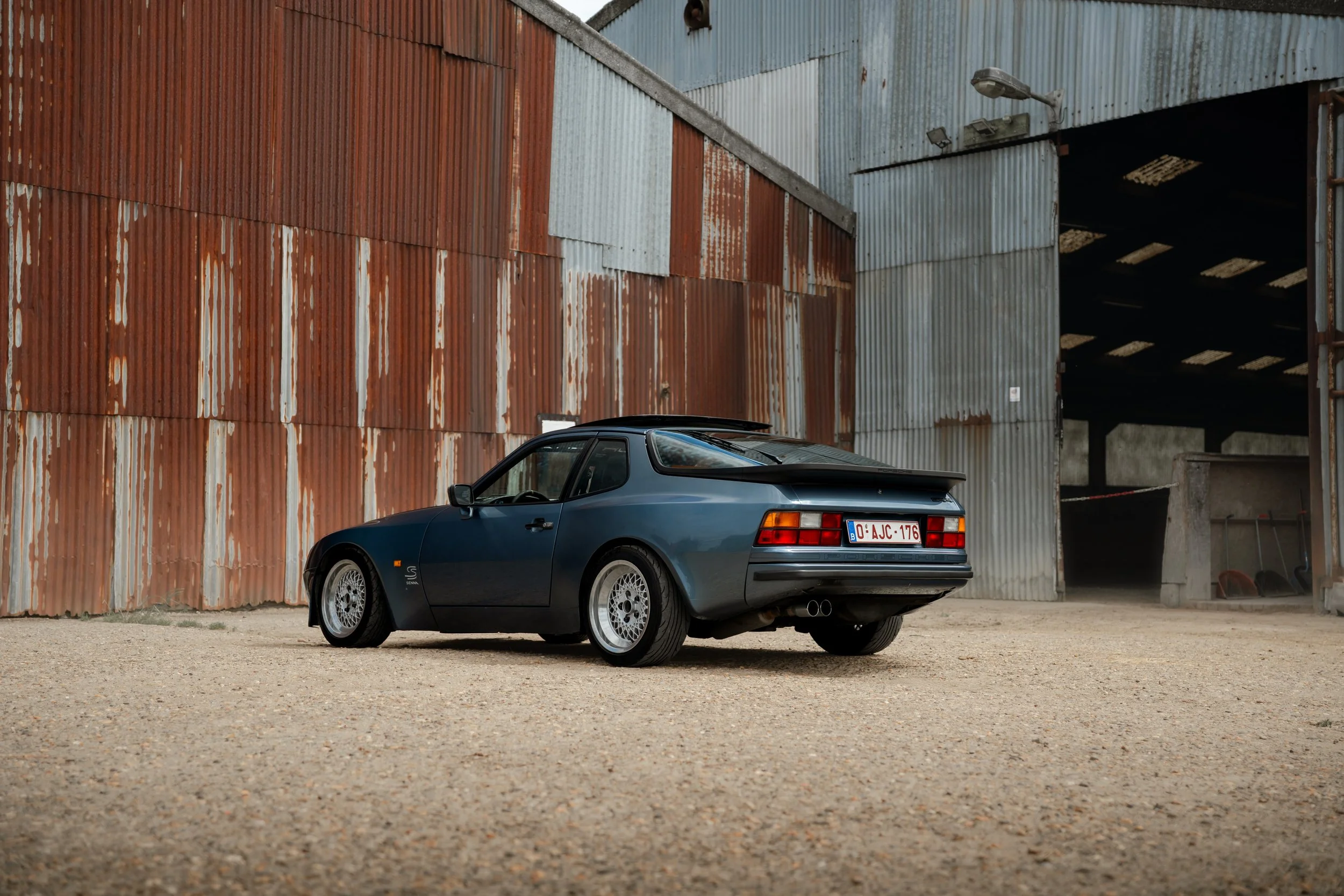 A blue Porsche sports car parked in front of a large, weathered, metal industrial building with rusted and corrugated panels.
