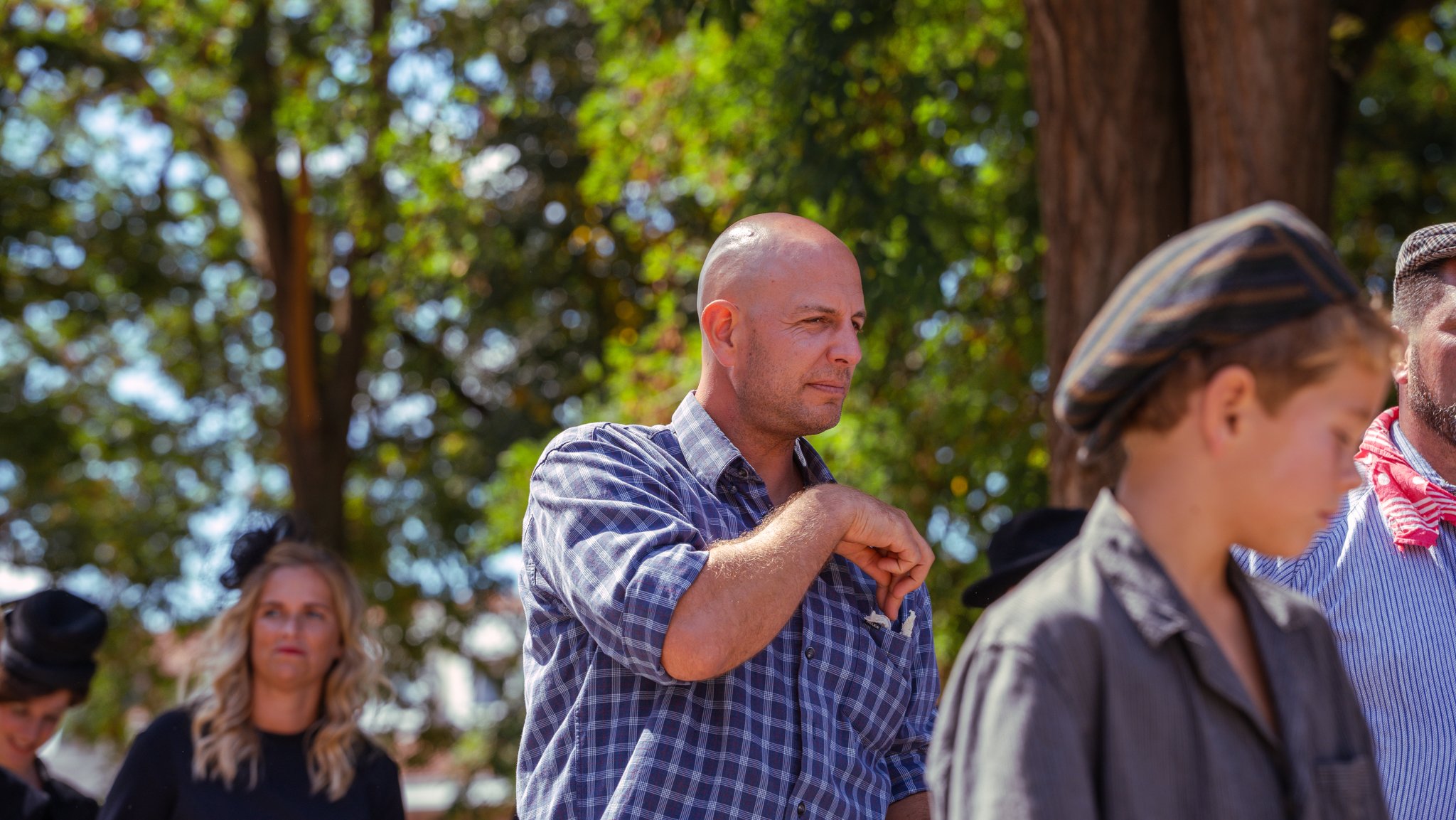 A group of people standing outdoors under trees, with one bald man in a checkered shirt folding his arms and looking thoughtful.