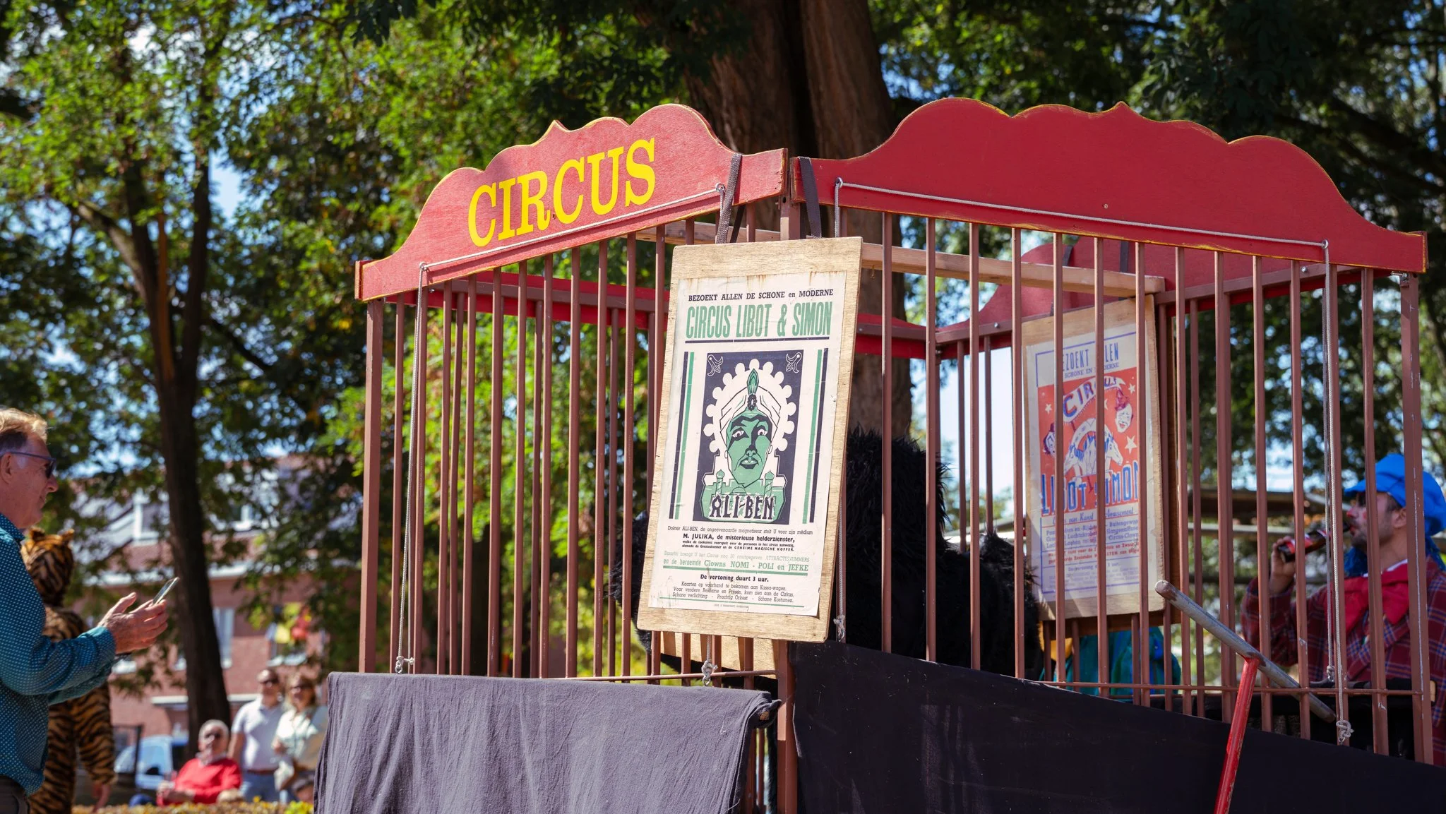 A small circus setup with a red sign that says 'CIRCUS' at the top. Inside, there are vintage posters advertising the circus, one of which features a green face with a headdress. There are people around the circus, including a person wearing a blue c