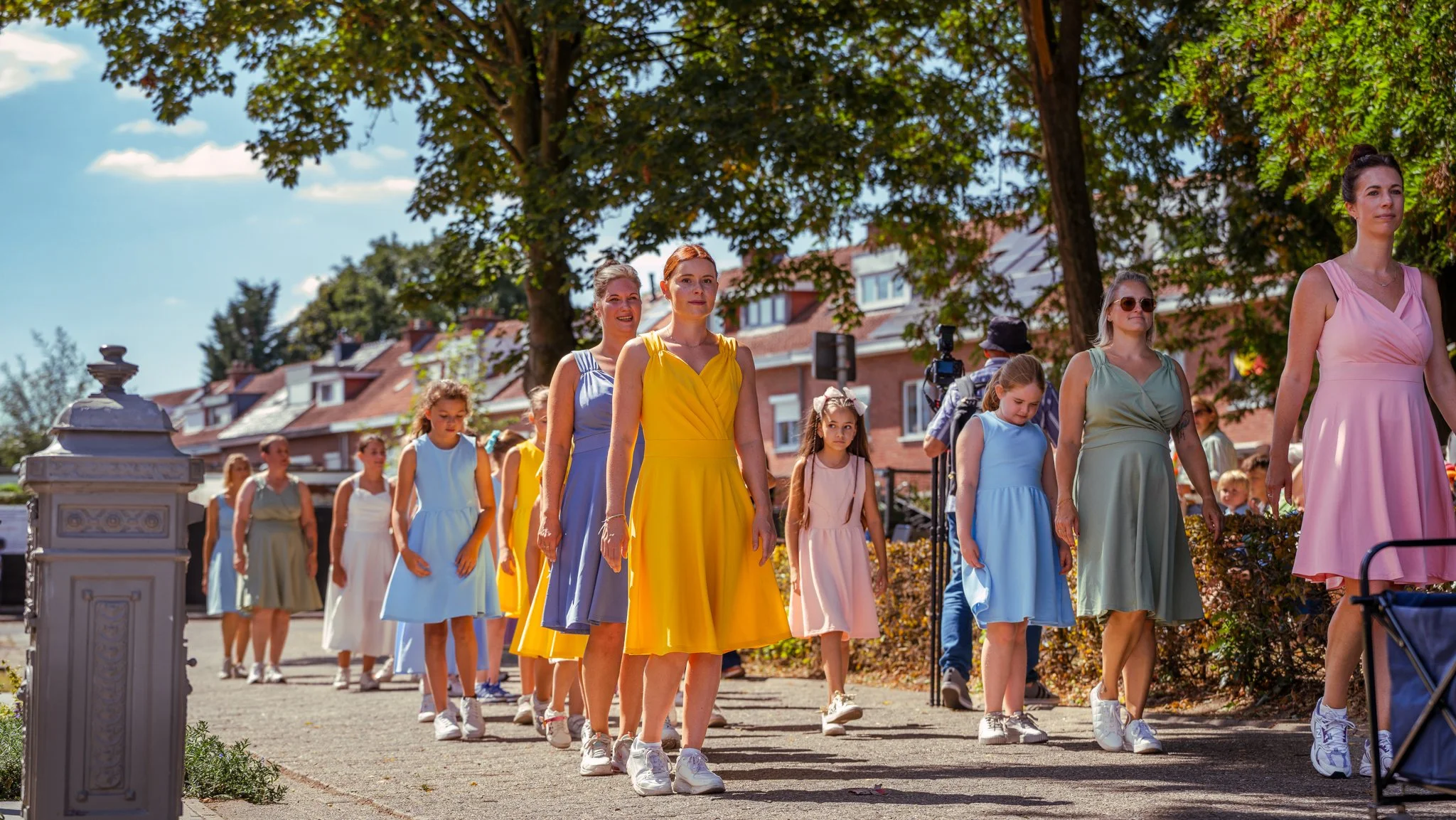 Women and girls walking outdoors in colorful dresses during daytime, with trees and houses in the background.