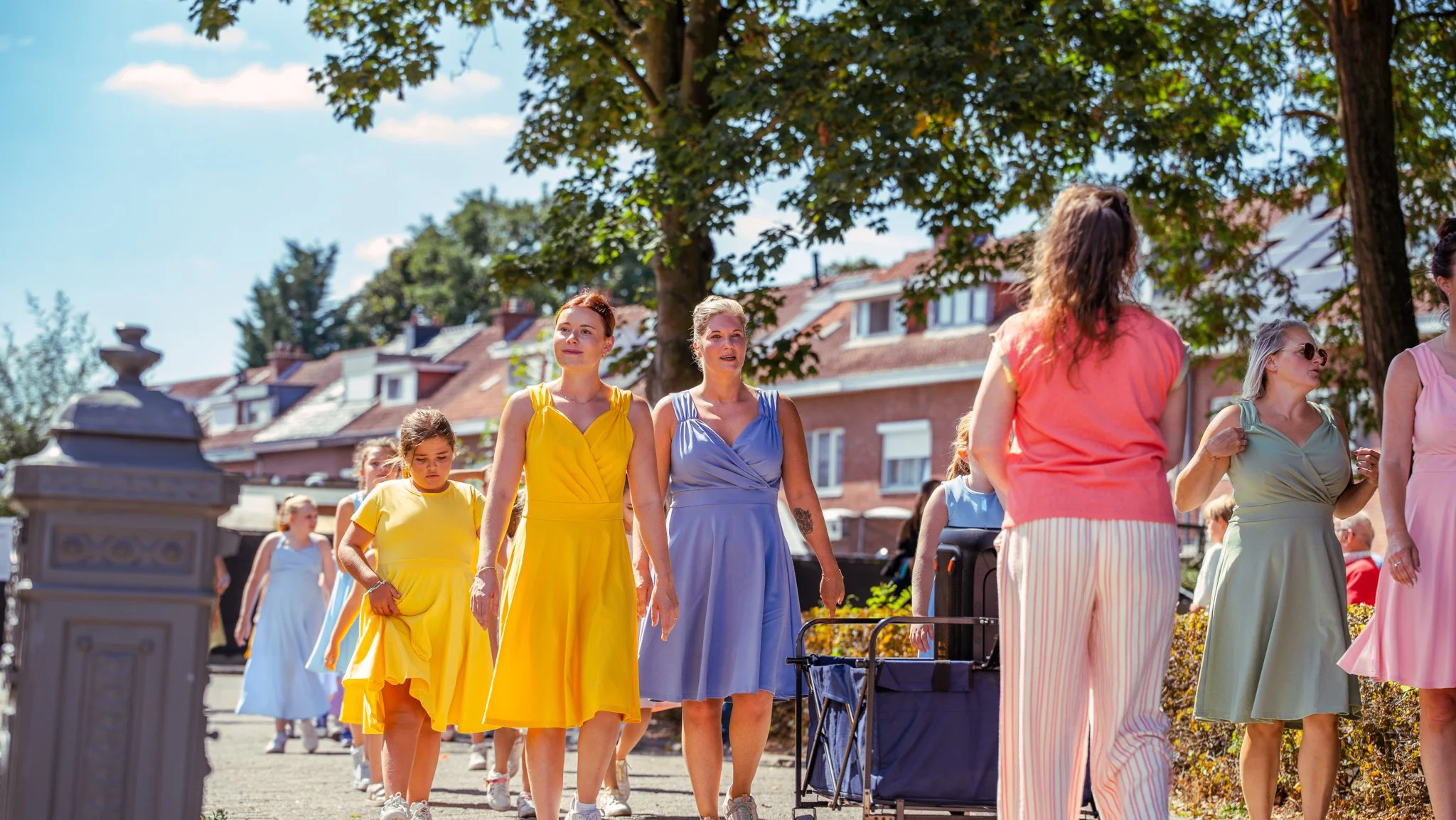 Group of women and girls walking outdoors on a sunny day, wearing colorful dresses, with trees and residential buildings in the background.