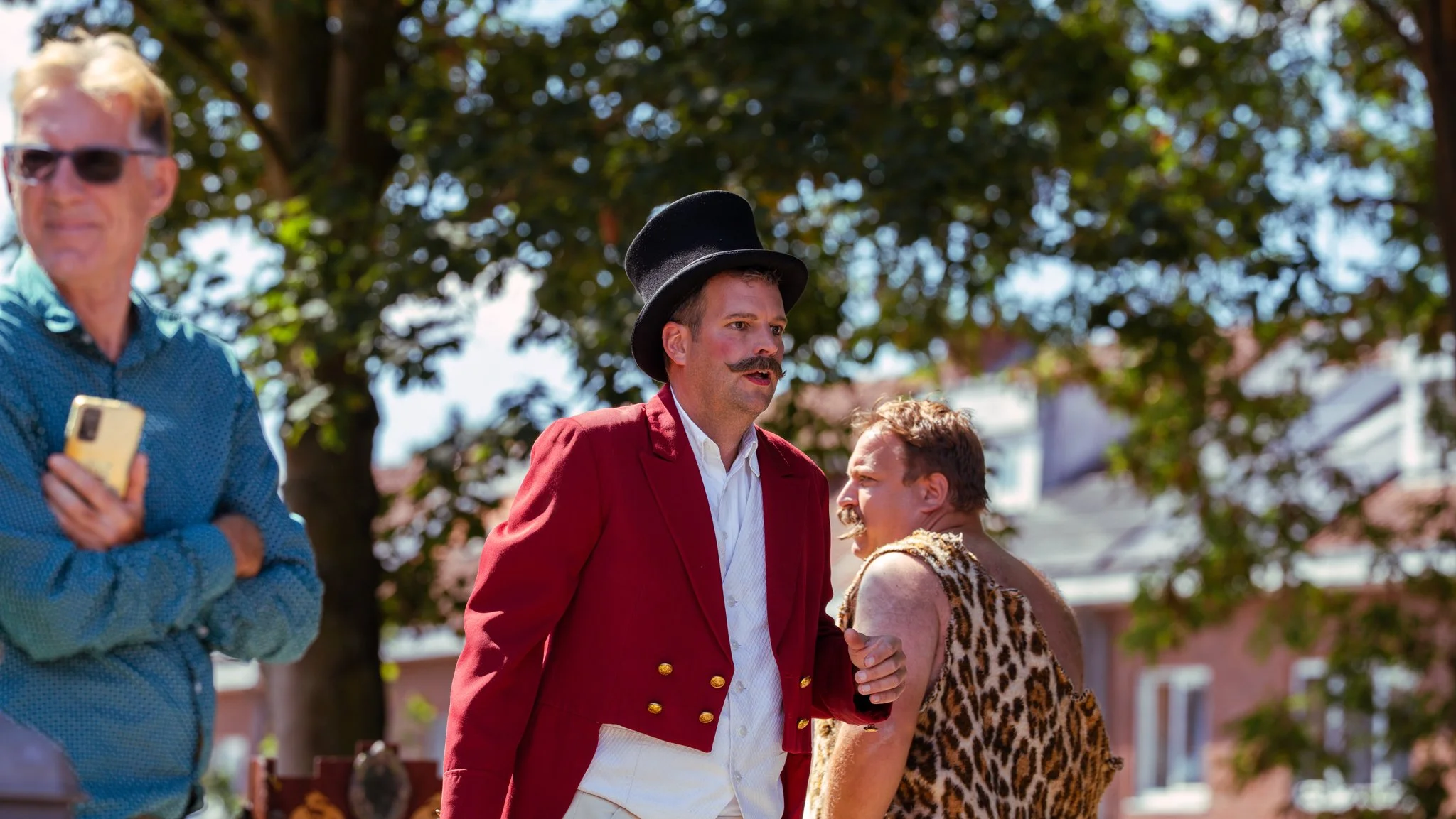 Three men outside among trees under a bright sky. The man in the center wears a red coat, white shirt, and a black top hat, with a mustache, and appears to be speaking or yelling. To his right, a man dons a leopard print sleeveless torso and has a sh