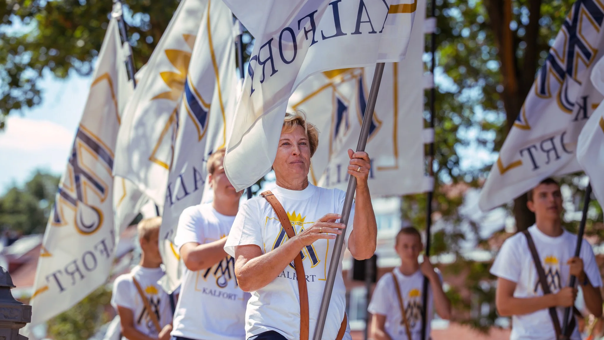 A group of people, led by a woman holding a large flag, participating in a parade or rally, wearing white T-shirts with a logo that includes a crown and the words 'California' and 'KALIFORNIA' visible, with flags bearing similar logos, under a sunny 