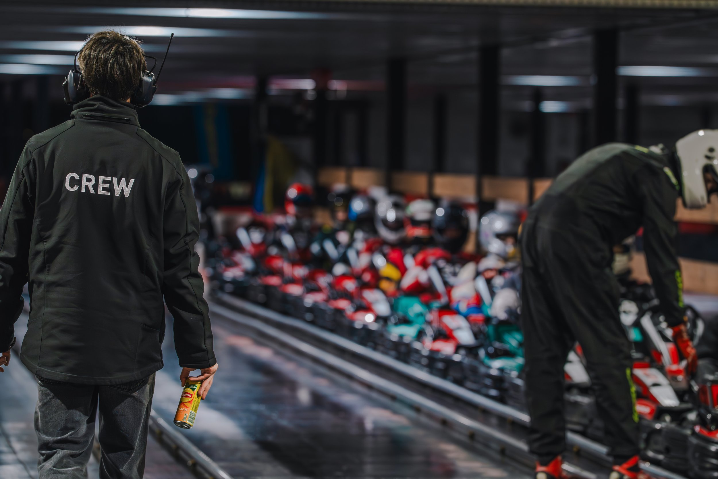 Person wearing black jacket with 'CREW' on back, holding a can of insect spray, walking past a display of helmets and sports gear in a warehouse or sporting goods store.