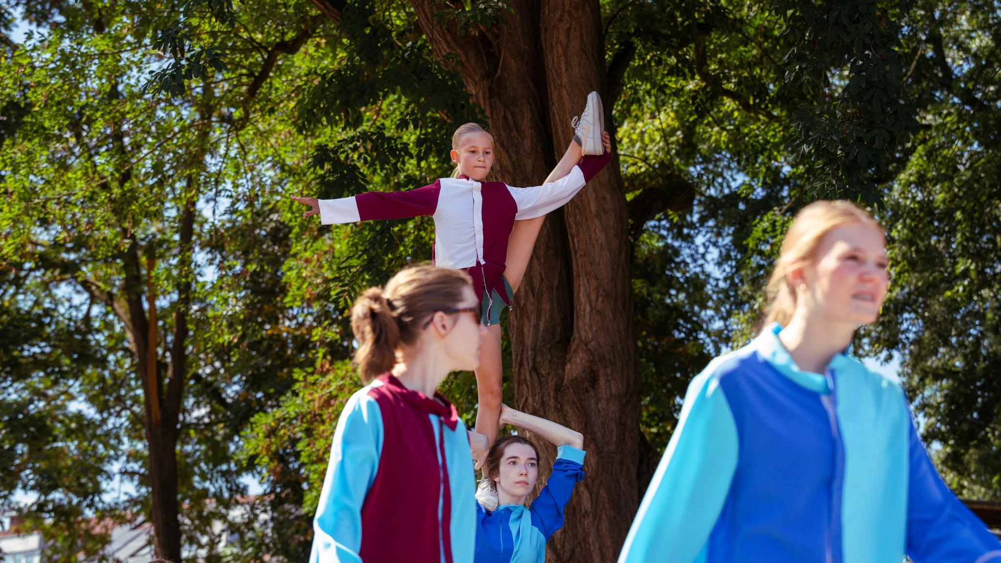 A girl in a maroon and white track jacket standing on a girl's hand, holding a split position near a large tree, with three other women in athletic jackets nearby, outdoors among green trees.