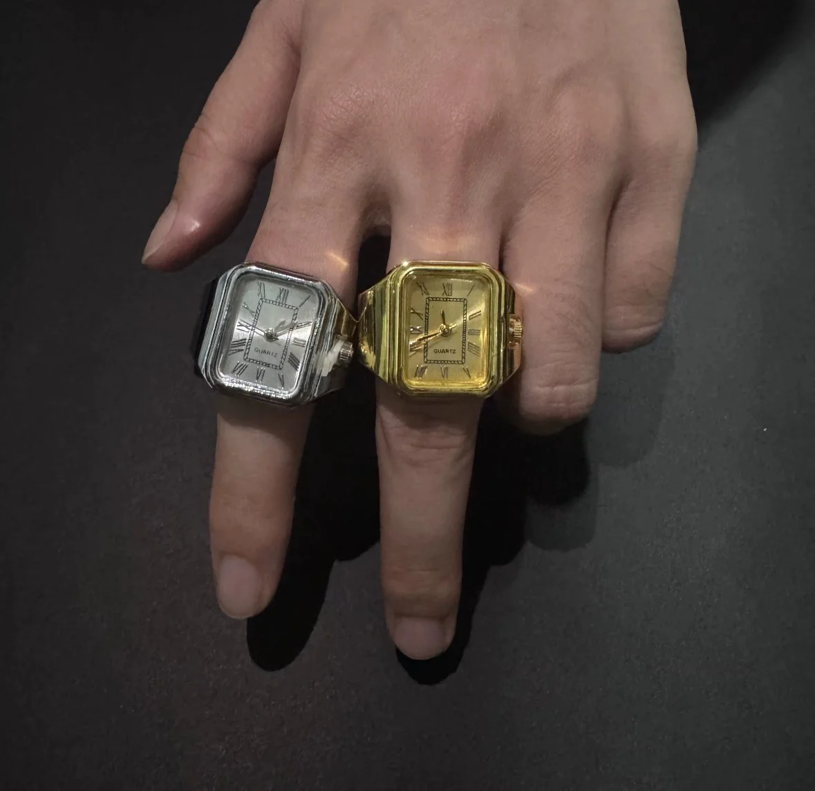 A hand wearing two large, square-shaped finger watch rings with rectangular faces; one watch is silver, and the other is gold, both with Roman numerals and black hands.