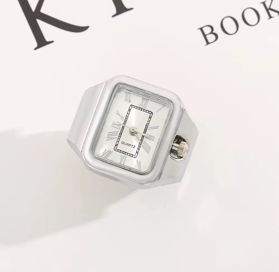 Silver square watch finger ring with a white face, Roman numerals, and a black rectangular date display, casting a shadow on a white surface.