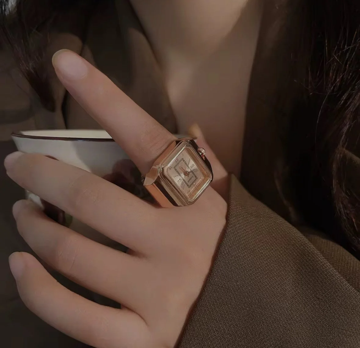 Close-up of a woman's hand with a large, square gold finger ring watch, holding a small teacup. She is wearing a brown suit jacket, and her dark hair is partially visible.