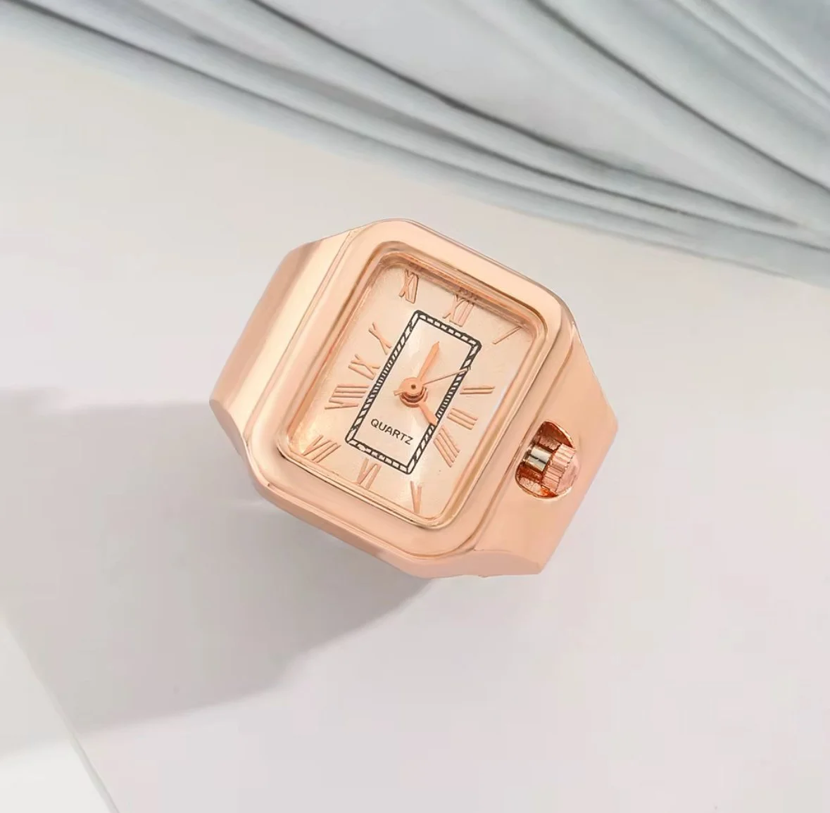 A pink quartz finger ring watch with a square face, Roman numerals, and a small crown on the side, placed on a white surface