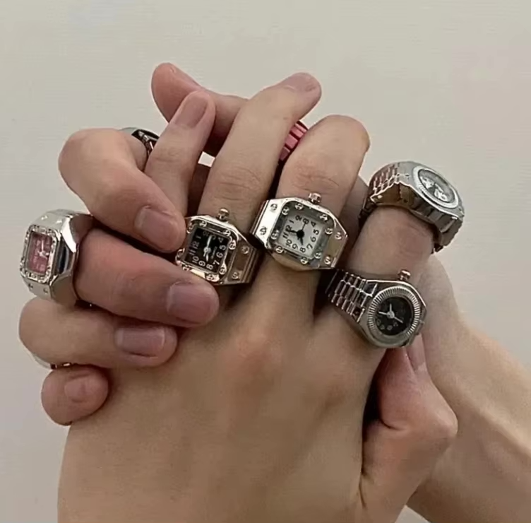 Dual fists in frame featuring a man’s hand and woman’s hand stacked with TinyTime ring watches: Pixie and Poem styles dominate, with crystal-rimmed bezels and colorful dials (pink, purple, black). Dramatic black outfit enhances the shine and detail o