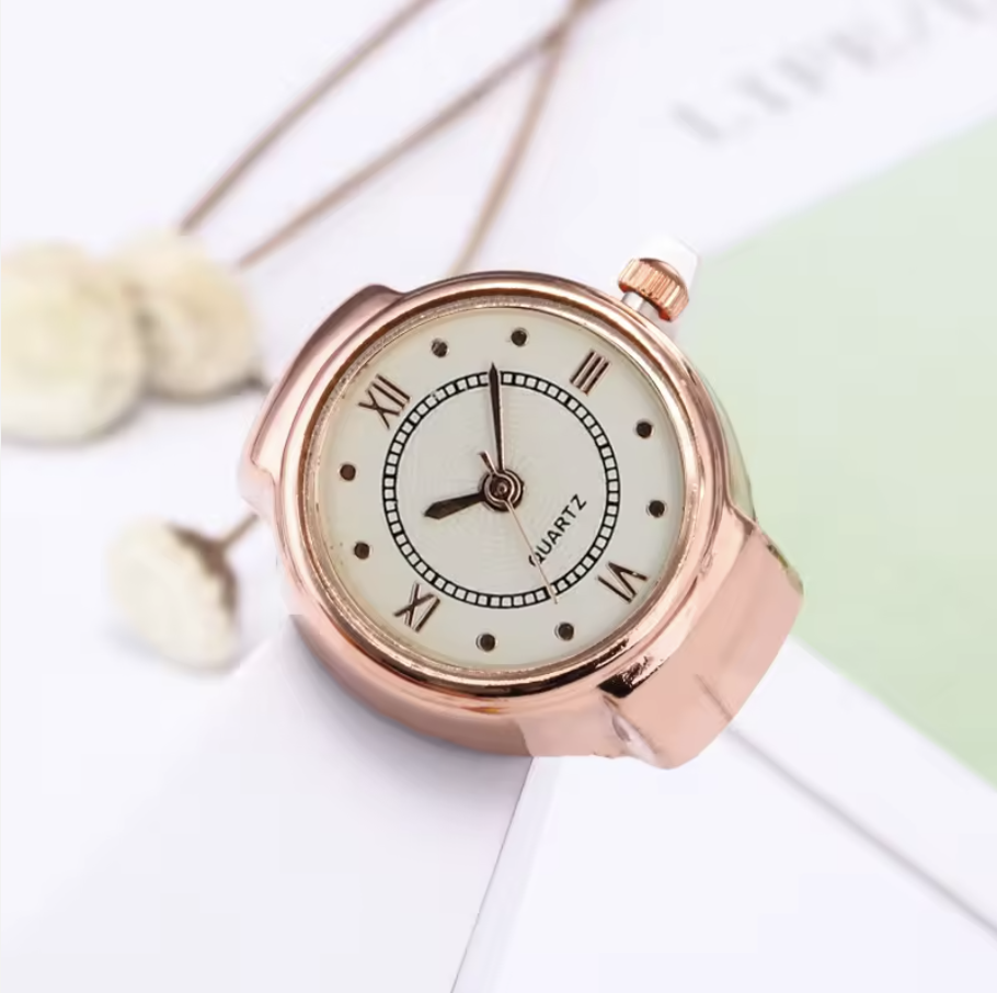 A rose gold ring watch MUSE with a white face, black hour and minute hands, and Roman numerals, resting on white paper with dried flowers nearby.