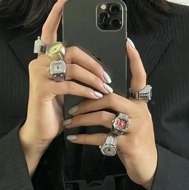 Woman holding a smartphone with both hands fully styled in TinyTime.co ring watches — Tinker, Pixie, Poem styles — in gold, silver, and bold red. A bold fashion statement merging utility with modern vintage design. Silver and gunmetal tones dominate 